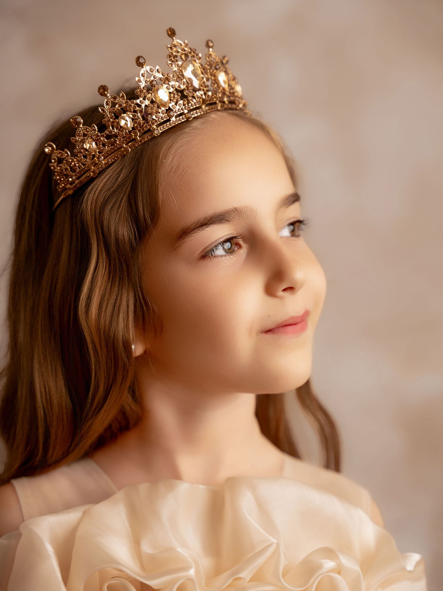 Young girl wearing an elegant gold crystal crown and cream dress, looking upward with a serene expression.
