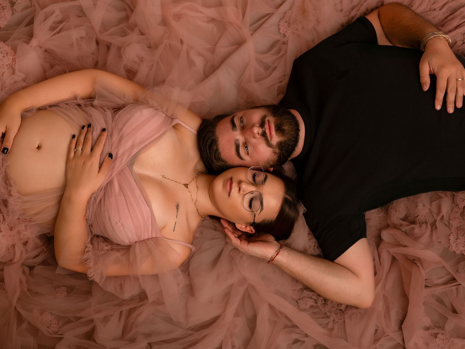 A pregnant couple lying on pink tulle fabric, the woman in a blush bralette cradling her bump as her partner embraces her.