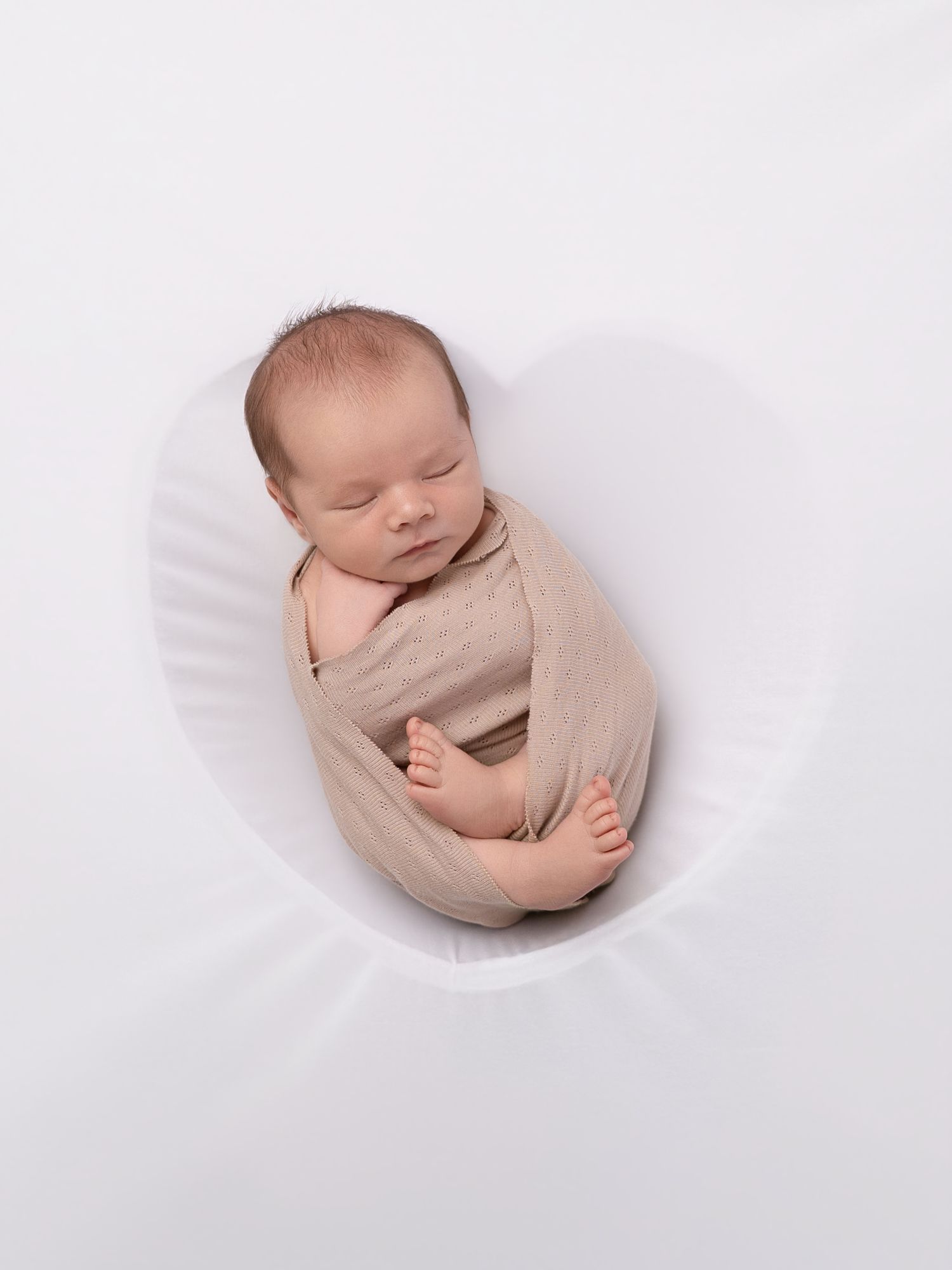 Sleeping newborn baby swaddled in a beige wrap, nestled in a white oval bowl for a studio photo.