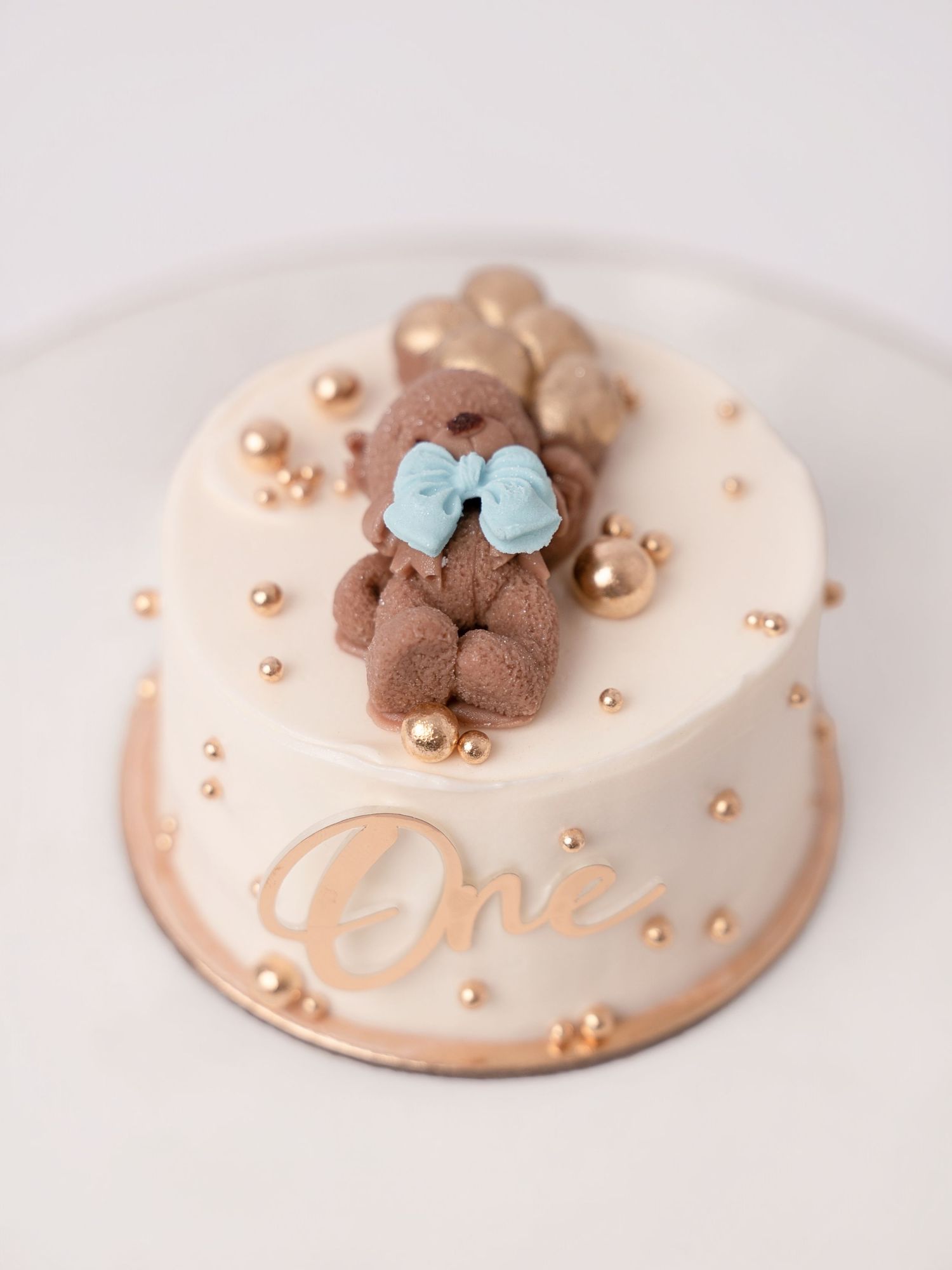 Elegant first birthday cake with teddy bear topper, blue bow, gold pearl decorations, and rose gold 'One' inscription.