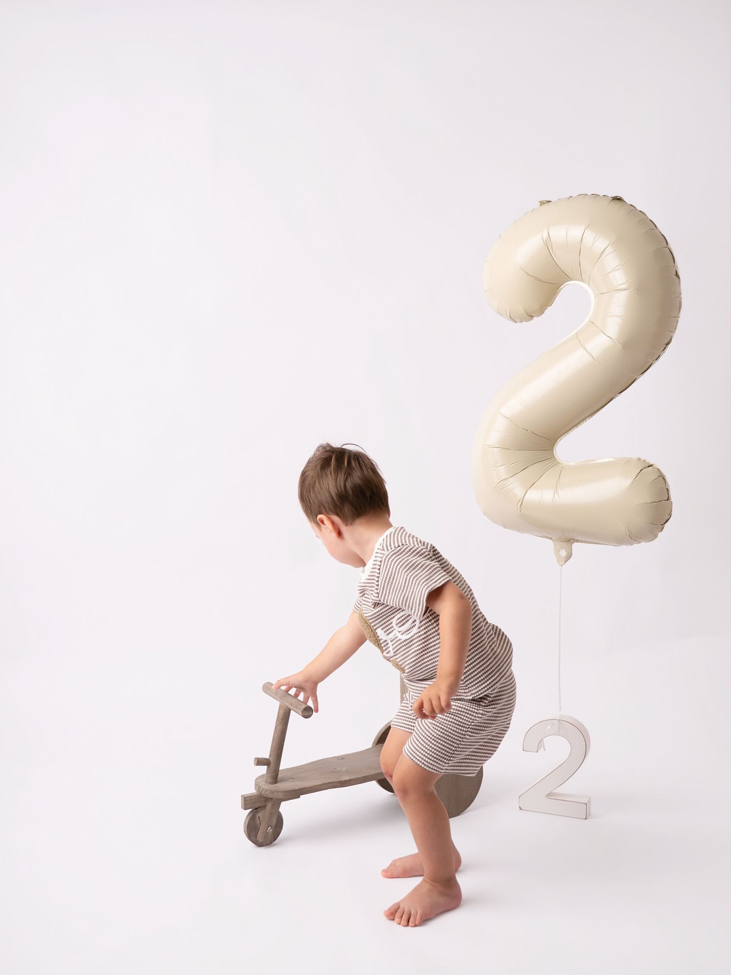 Toddler boy playing with wooden scooter beside number 2 balloon and decoration, celebrating 2nd birthday.