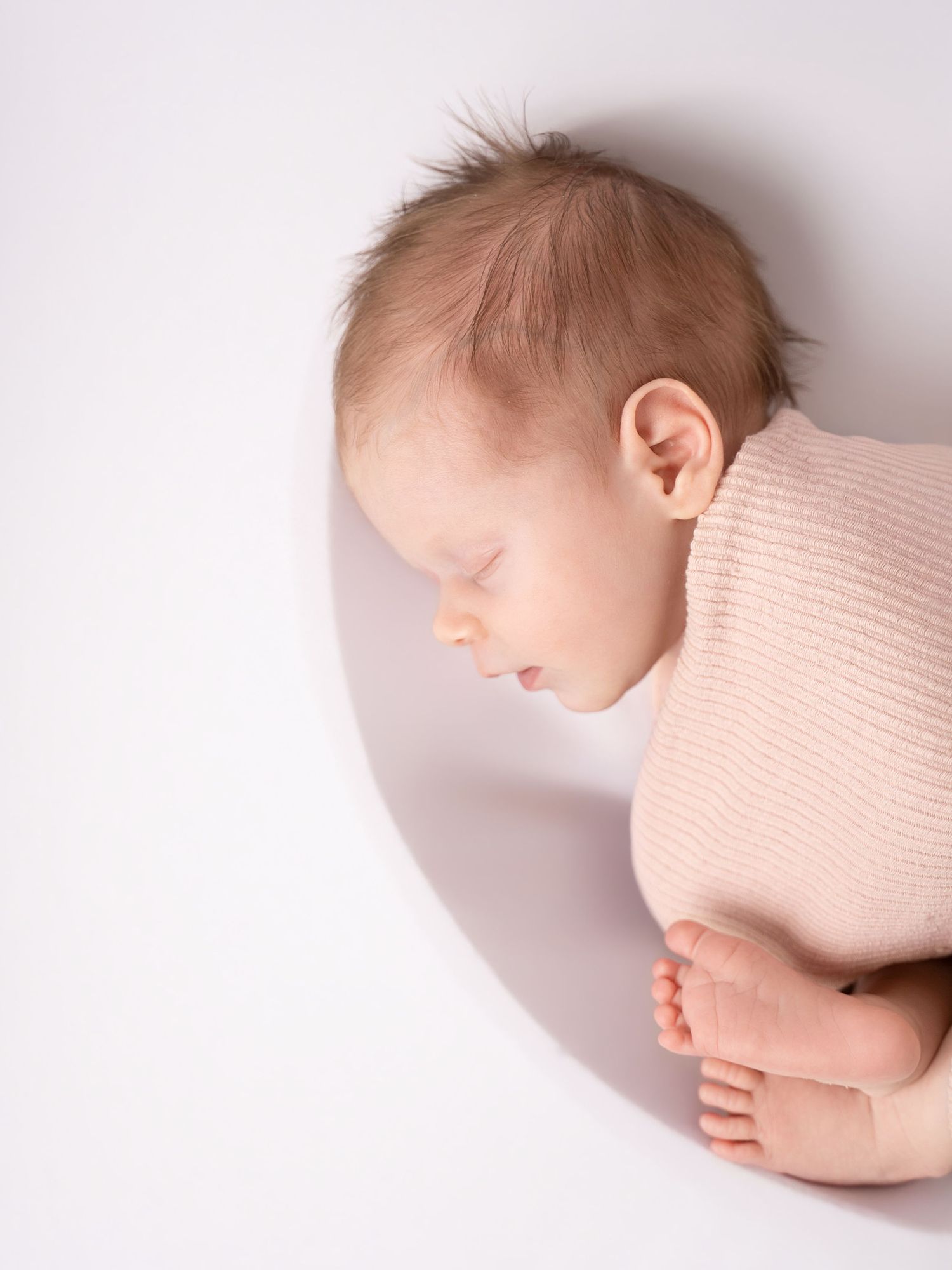 A peaceful newborn baby in a pink ribbed outfit sleeps curled up on a white background, showing tiny toes.