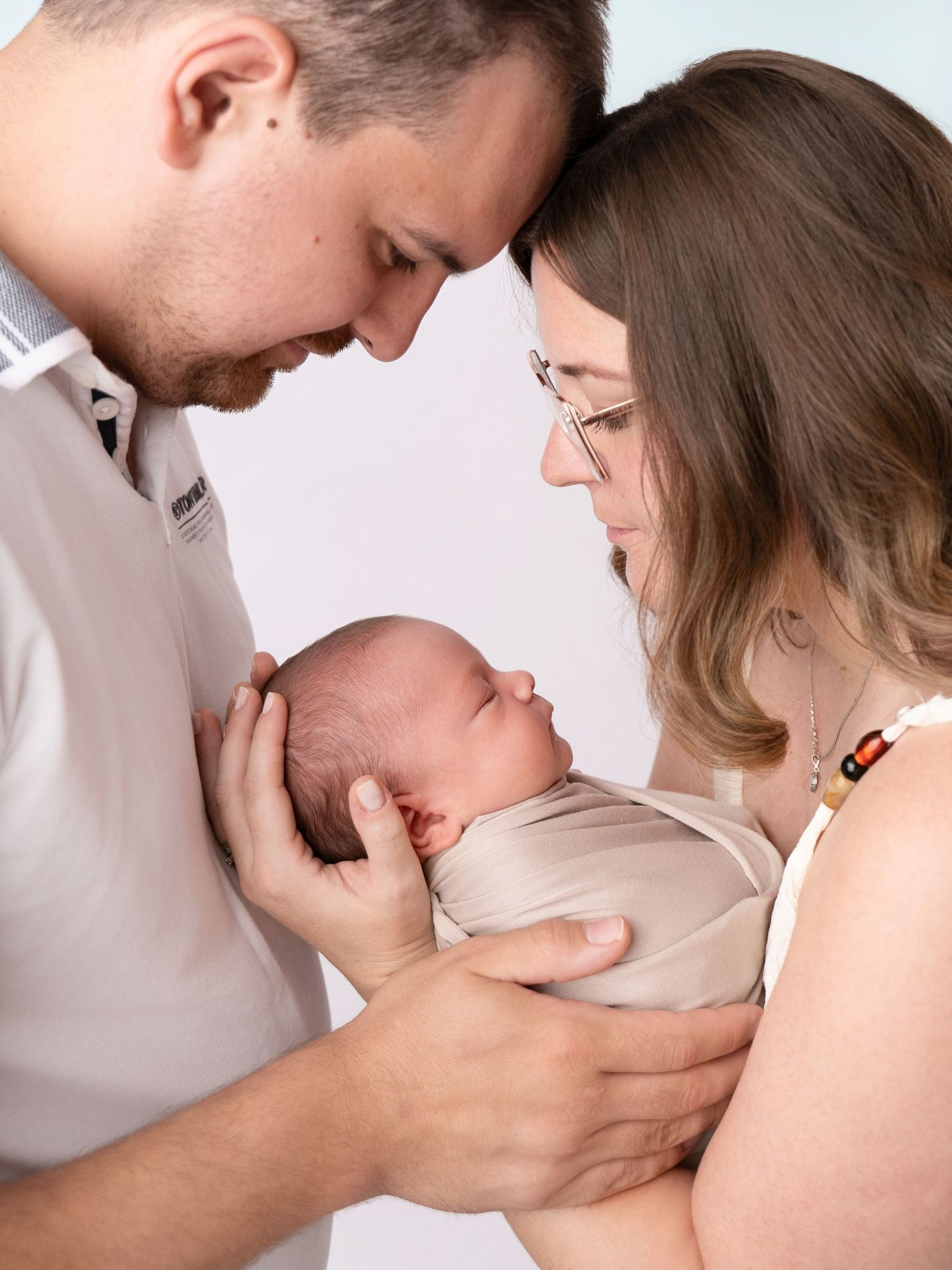 A couple tenderly holds their newborn baby, leaning in close together in a soft studio portrait.