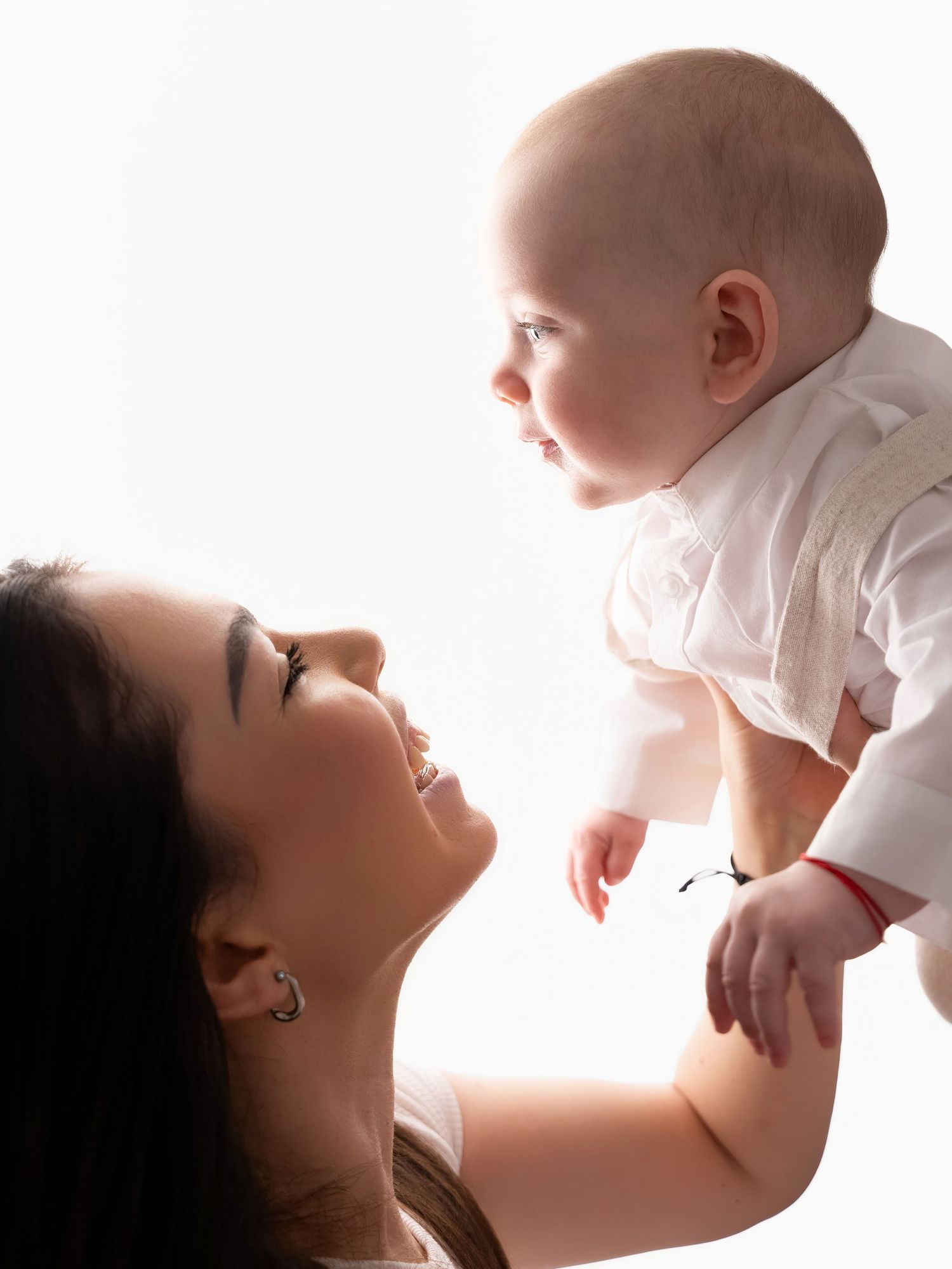 A smiling mother lifts her happy baby in the air against a bright white background.