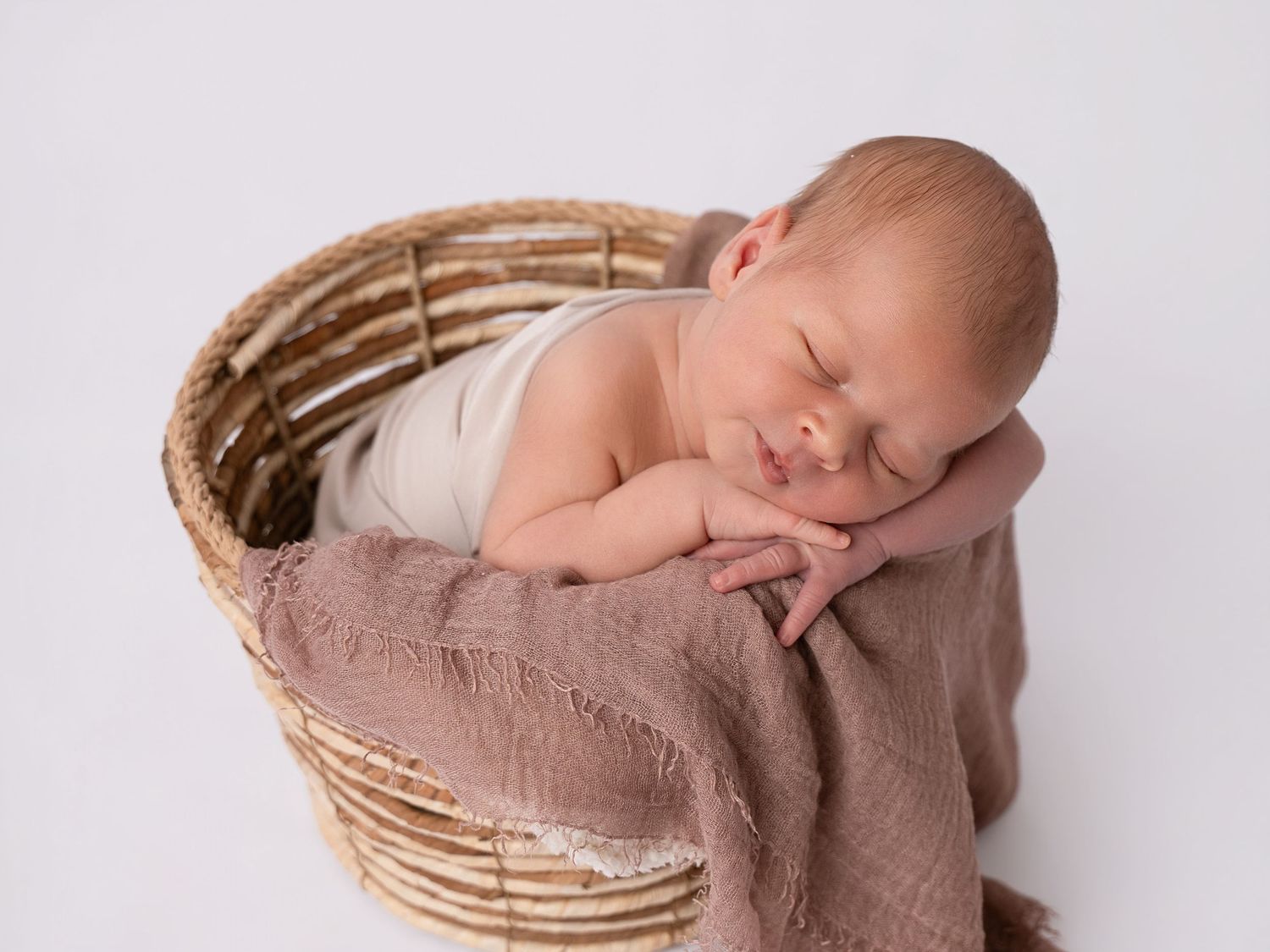 Peaceful newborn baby sleeping in a wicker basket wrapped in a soft mauve blanket on a white background.