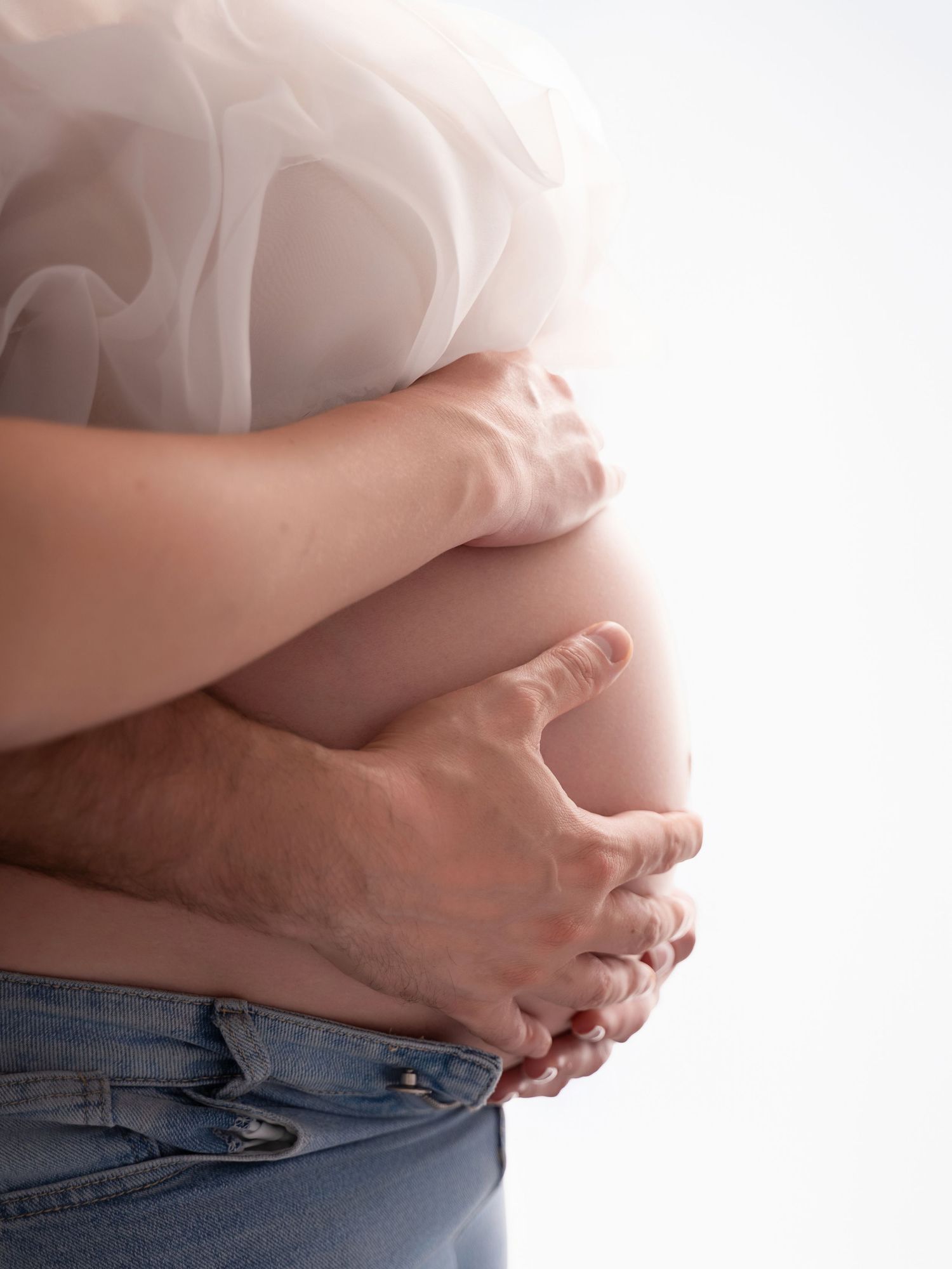 Couple embracing pregnant belly, man's hands gently cradling baby bump, maternity photography on white background.