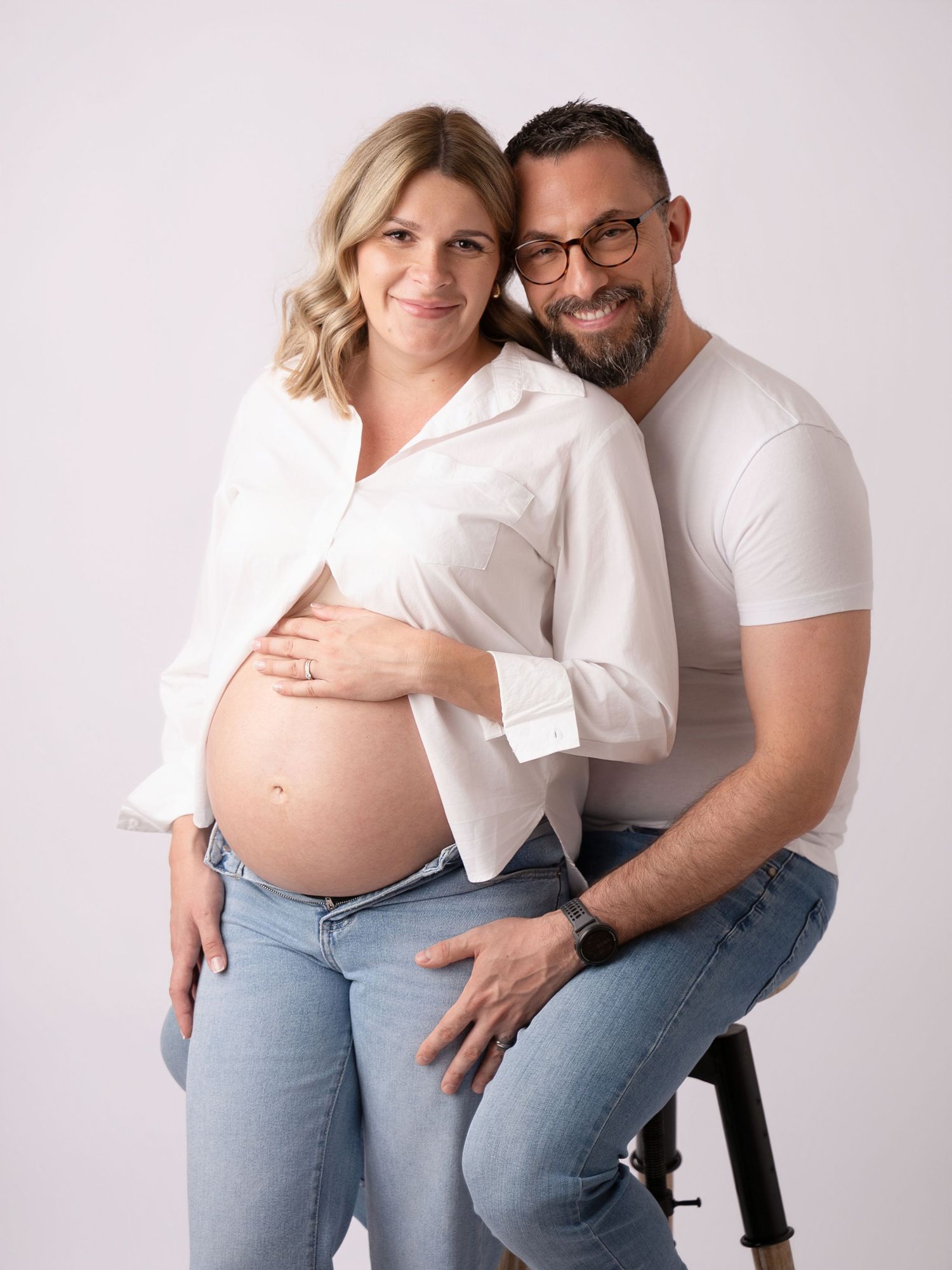 Couple in white shirts and jeans posing for maternity photo, man embracing pregnant woman showing baby bump.