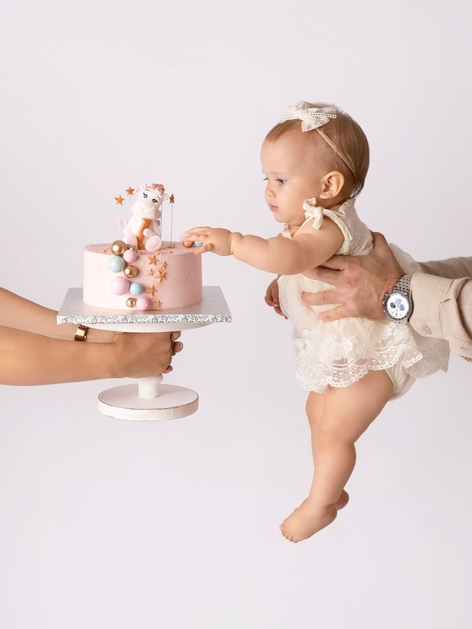A baby in a lace romper reaches for a pink first birthday cake held by an adult on a white background.