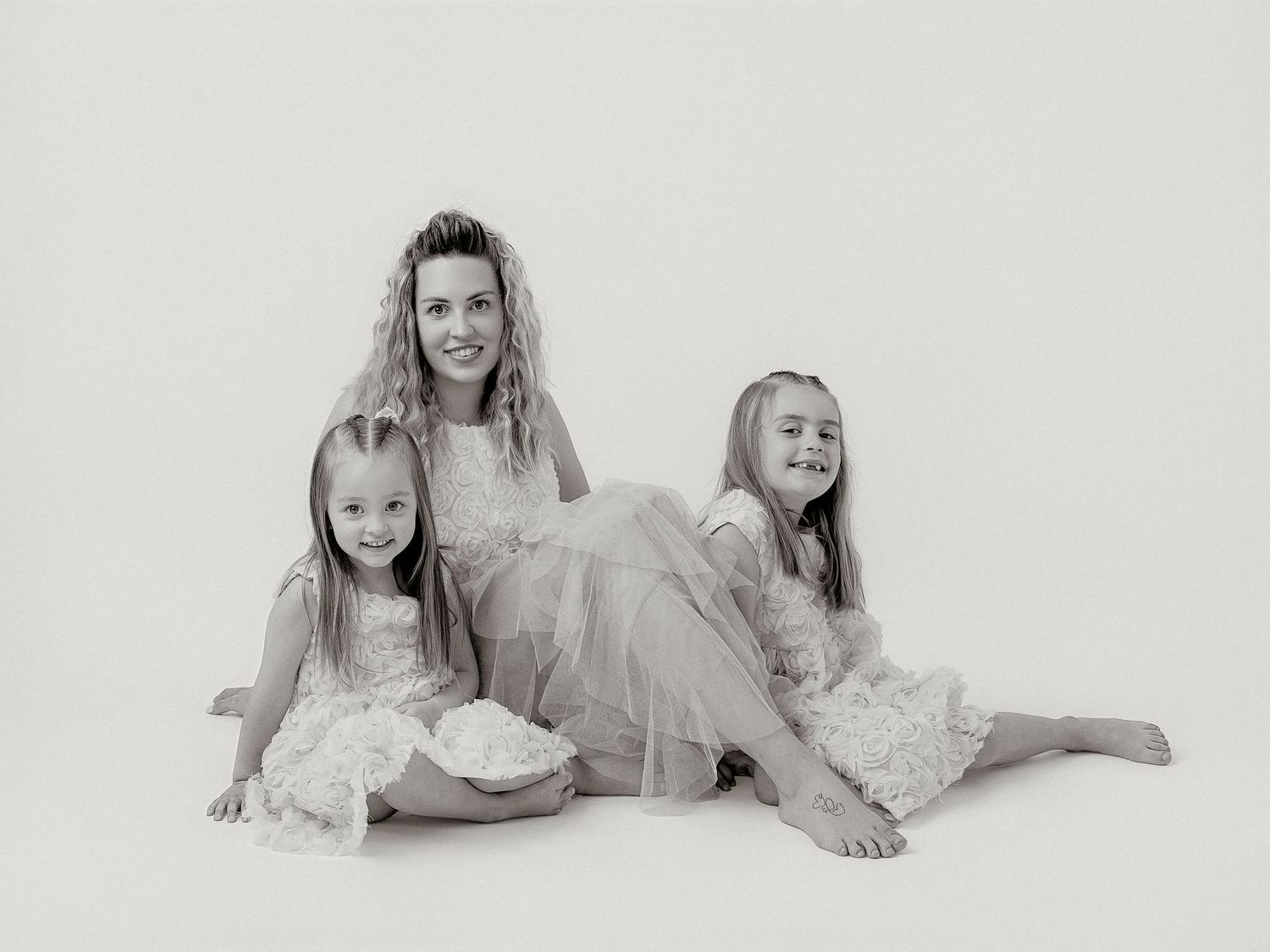 Black and white studio portrait of a mother and two young daughters in elegant dresses, posing together on white background.