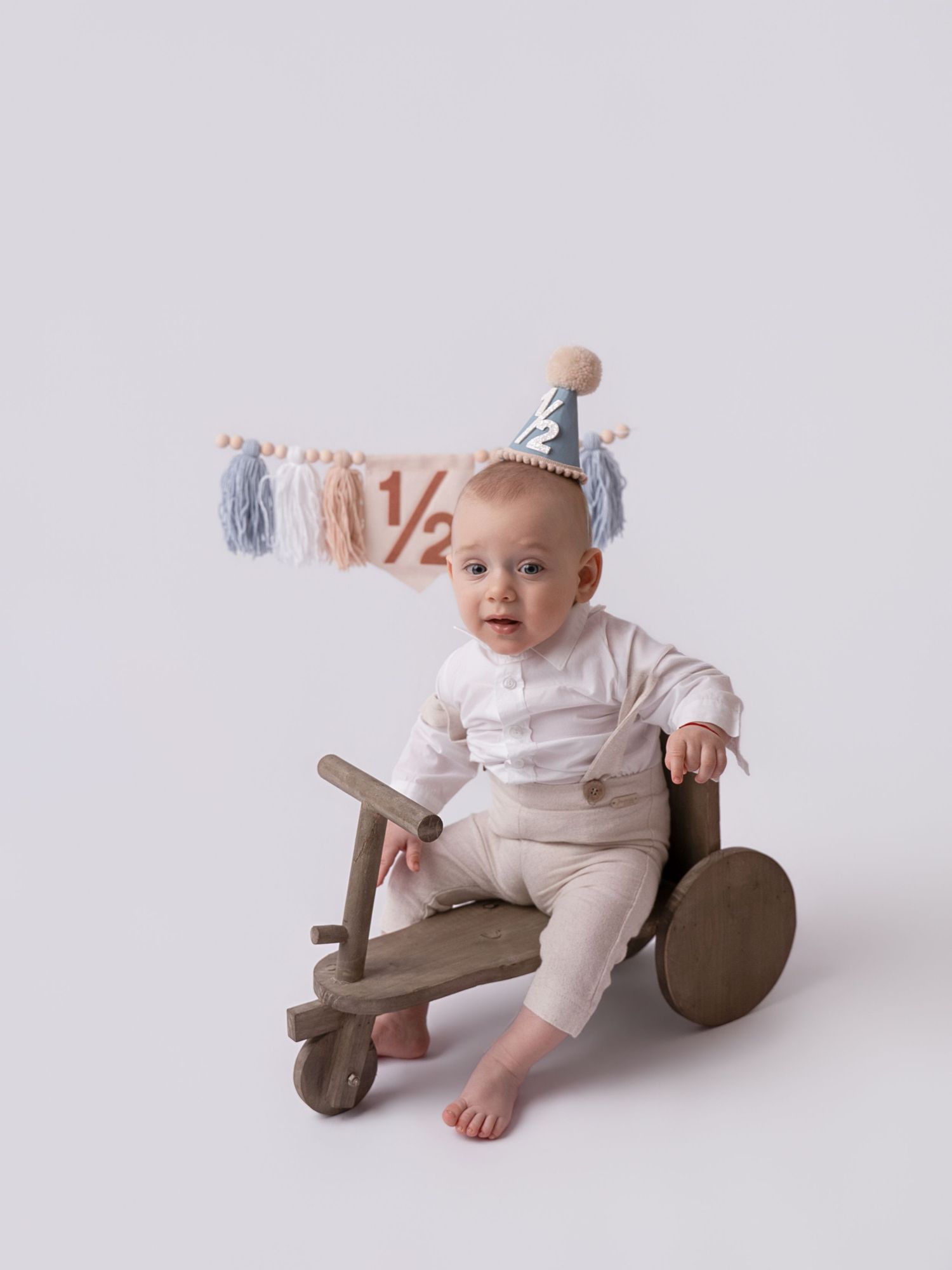 Baby in white outfit sits on wooden toy wearing a half birthday party hat with a 1/2 banner behind them.