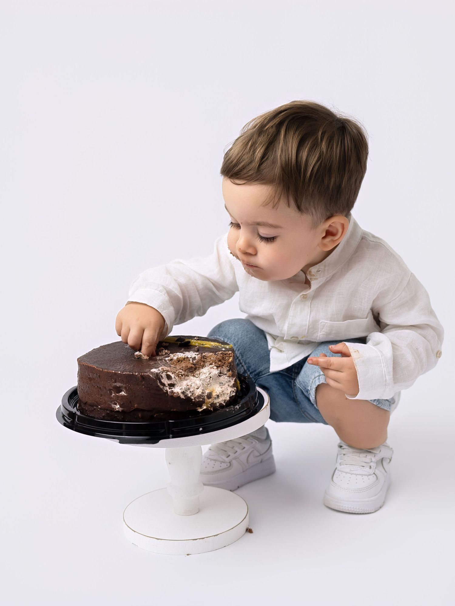 Toddler boy in white shirt and jeans reaching into a chocolate cake on a white pedestal stand.