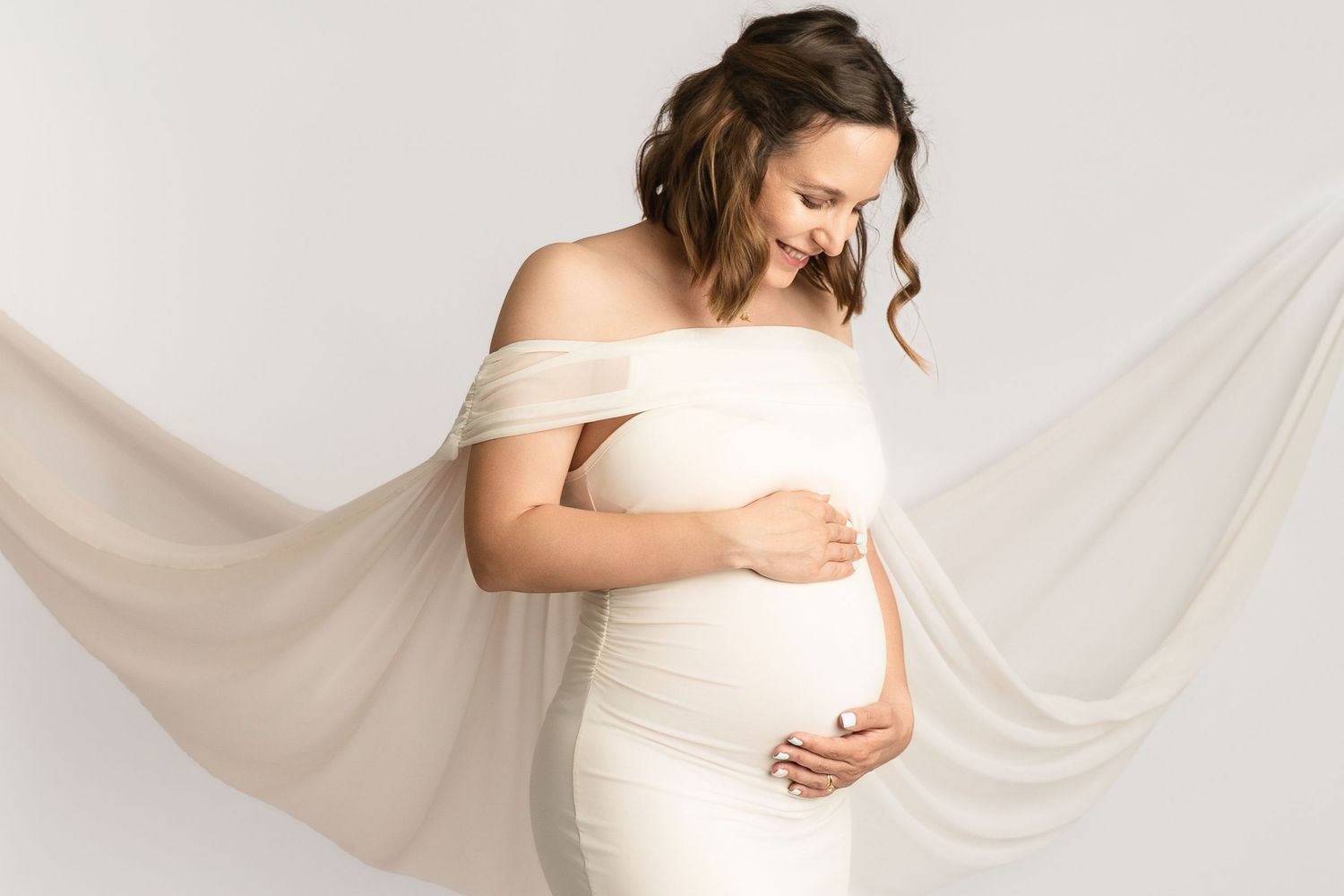 Pregnant woman in white off-shoulder dress holding baby bump with flowing fabric drape in studio.