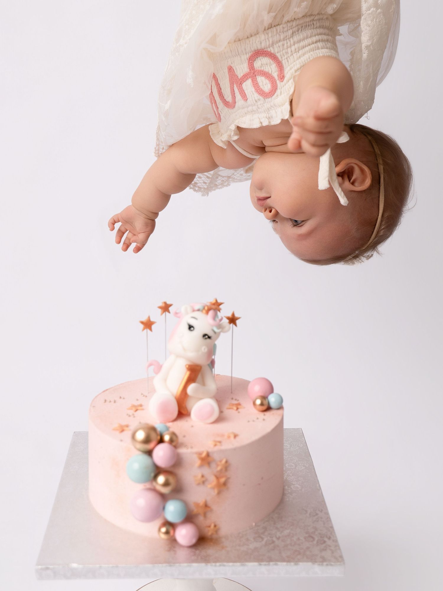 Baby girl leans over pink unicorn first birthday smash cake with pastel balloon decorations.