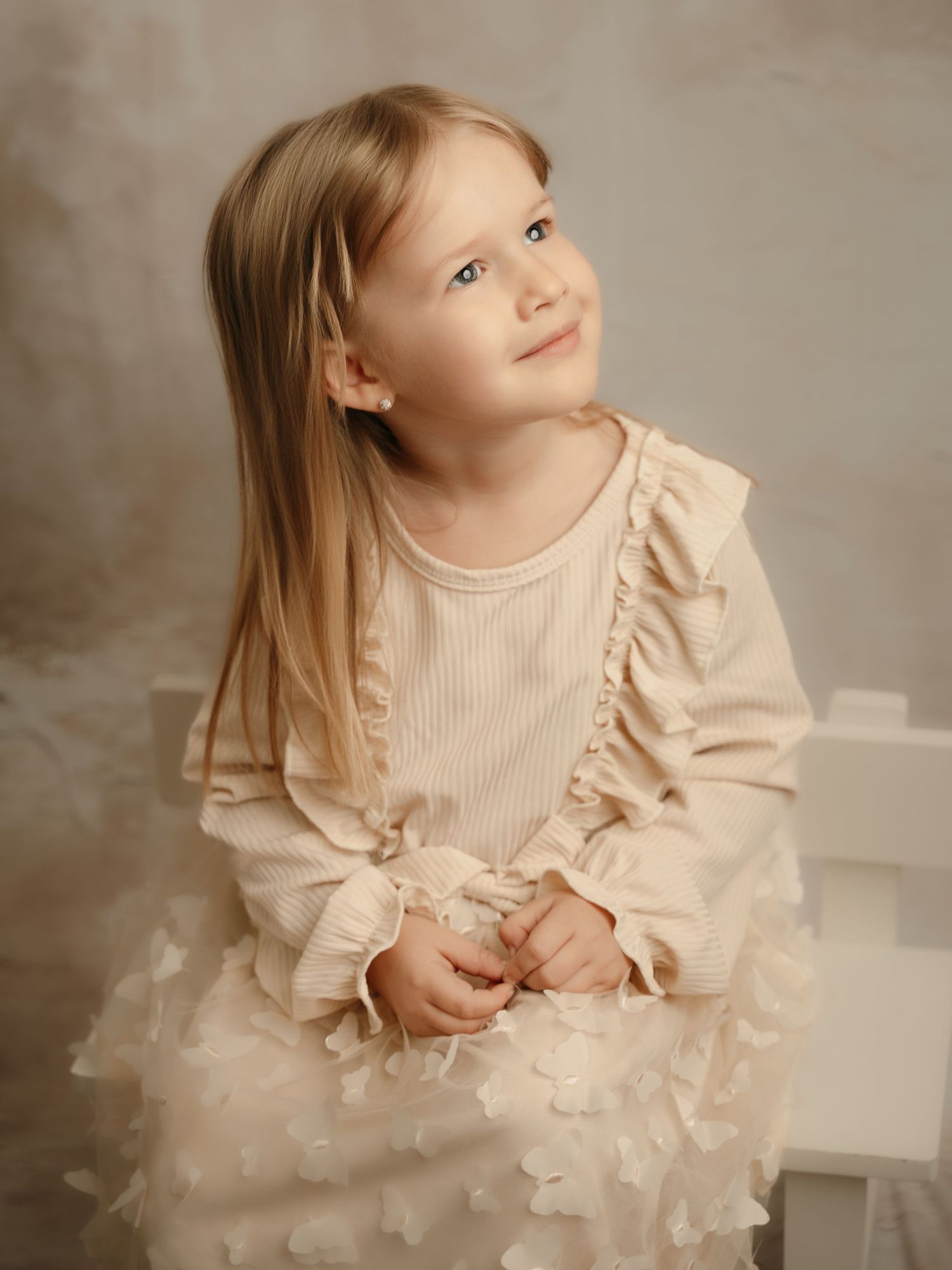 Blonde girl in a cream ribbed dress with ruffles and floral tulle skirt, sitting and gazing upward softly.