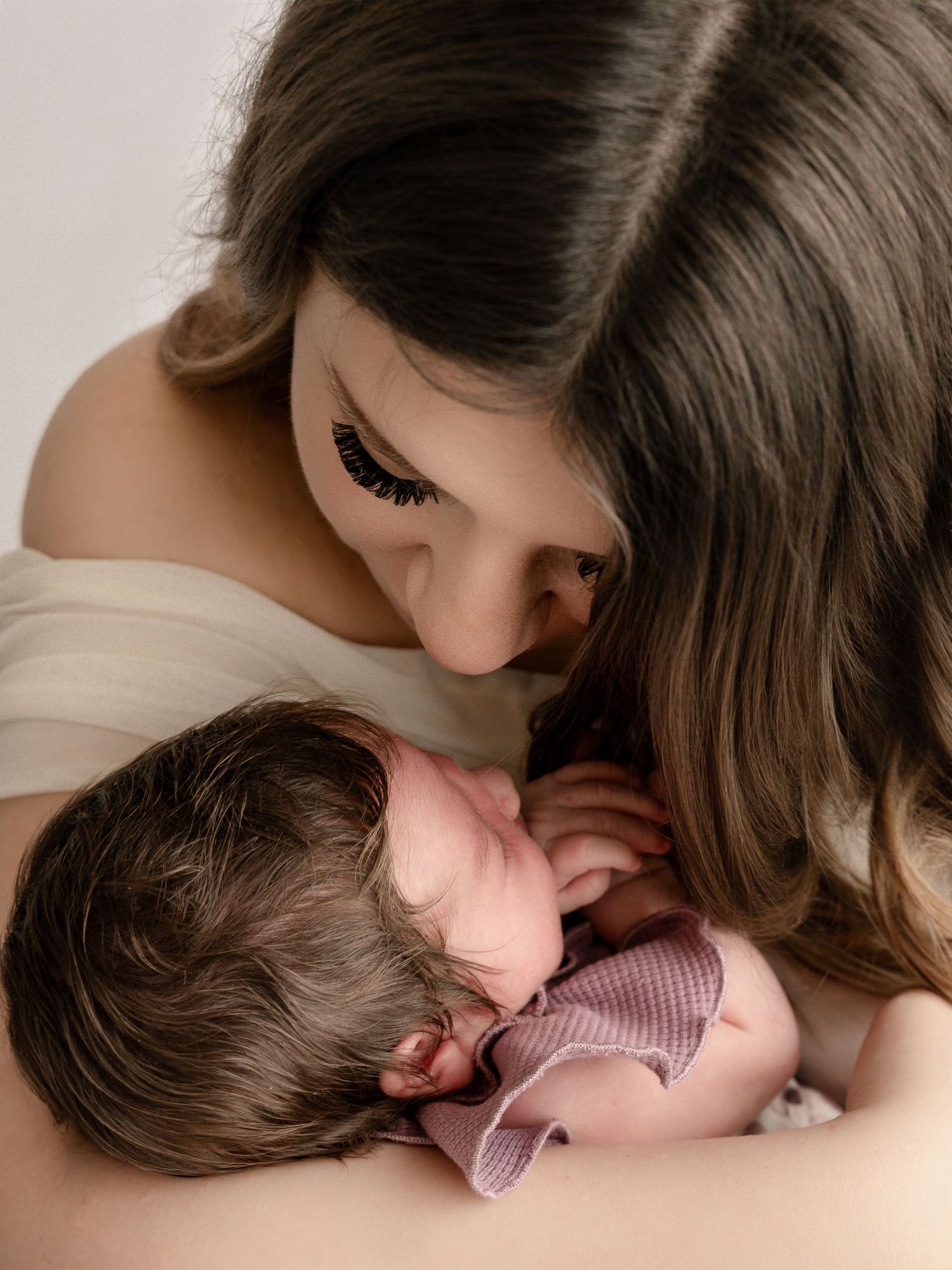 Loving mother with long brown hair gazes down tenderly at sleeping newborn baby in pink outfit.