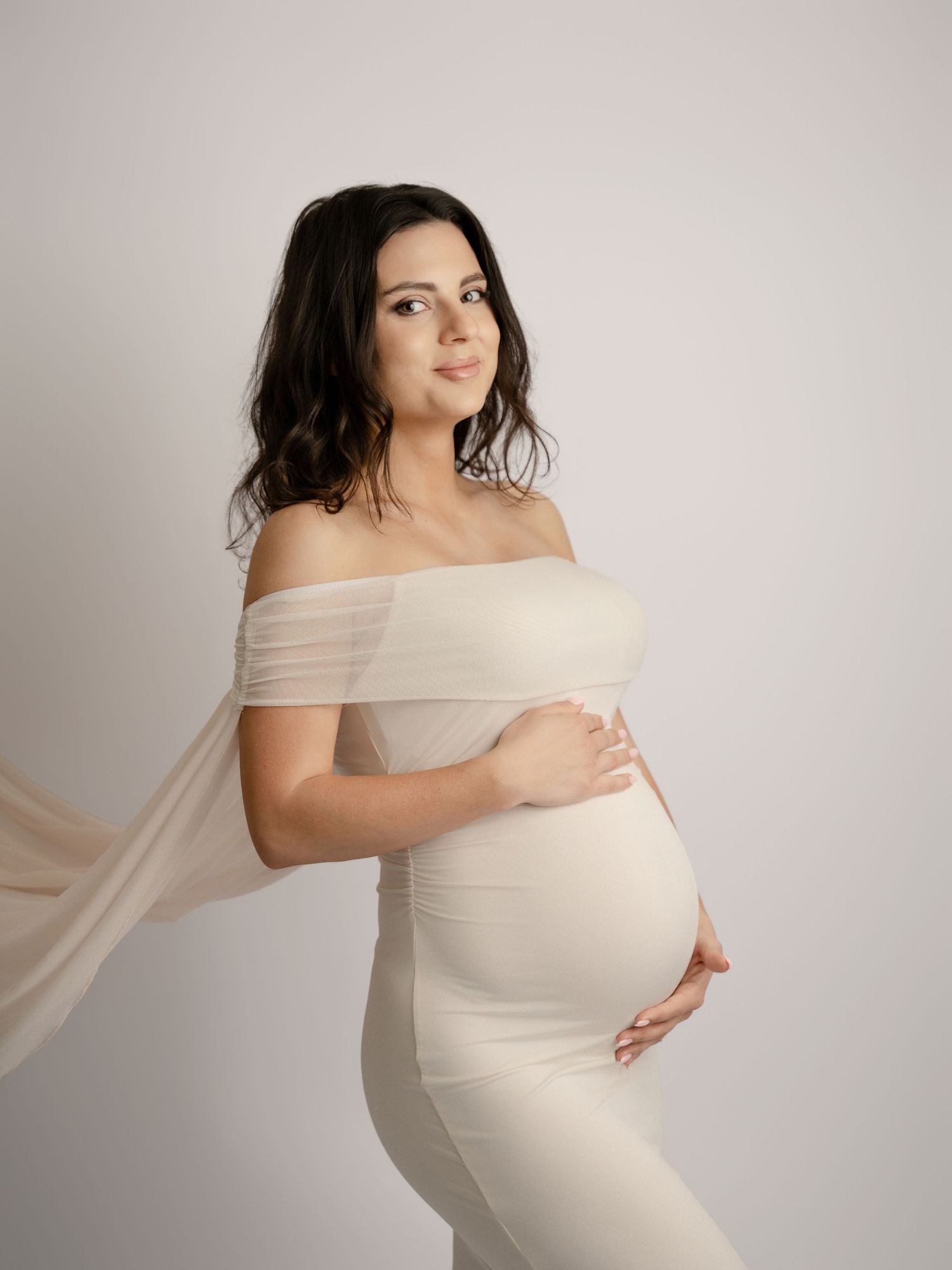 Pregnant woman in cream off-shoulder dress gently cradles baby bump in soft studio portrait.