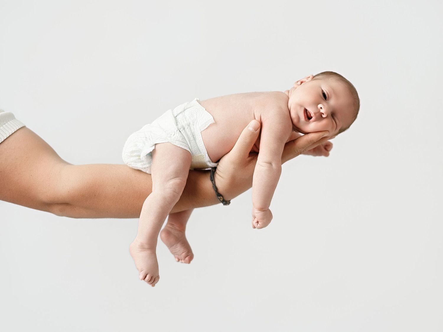 Adult hand carefully supporting a smiling newborn baby in a diaper on white background.