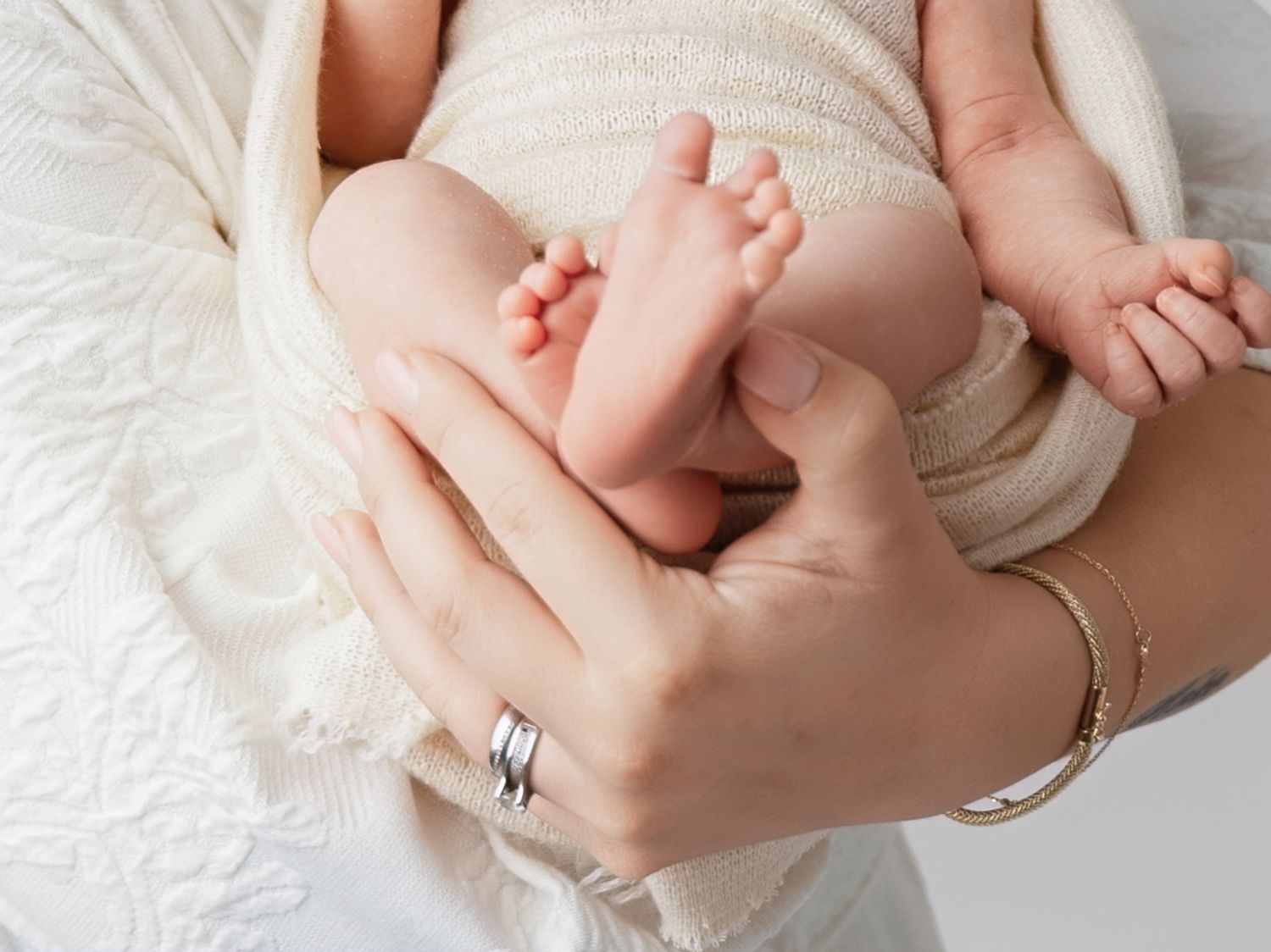 A mother gently cradles her newborn baby wrapped in a soft cream blanket, showing tiny feet and hands.