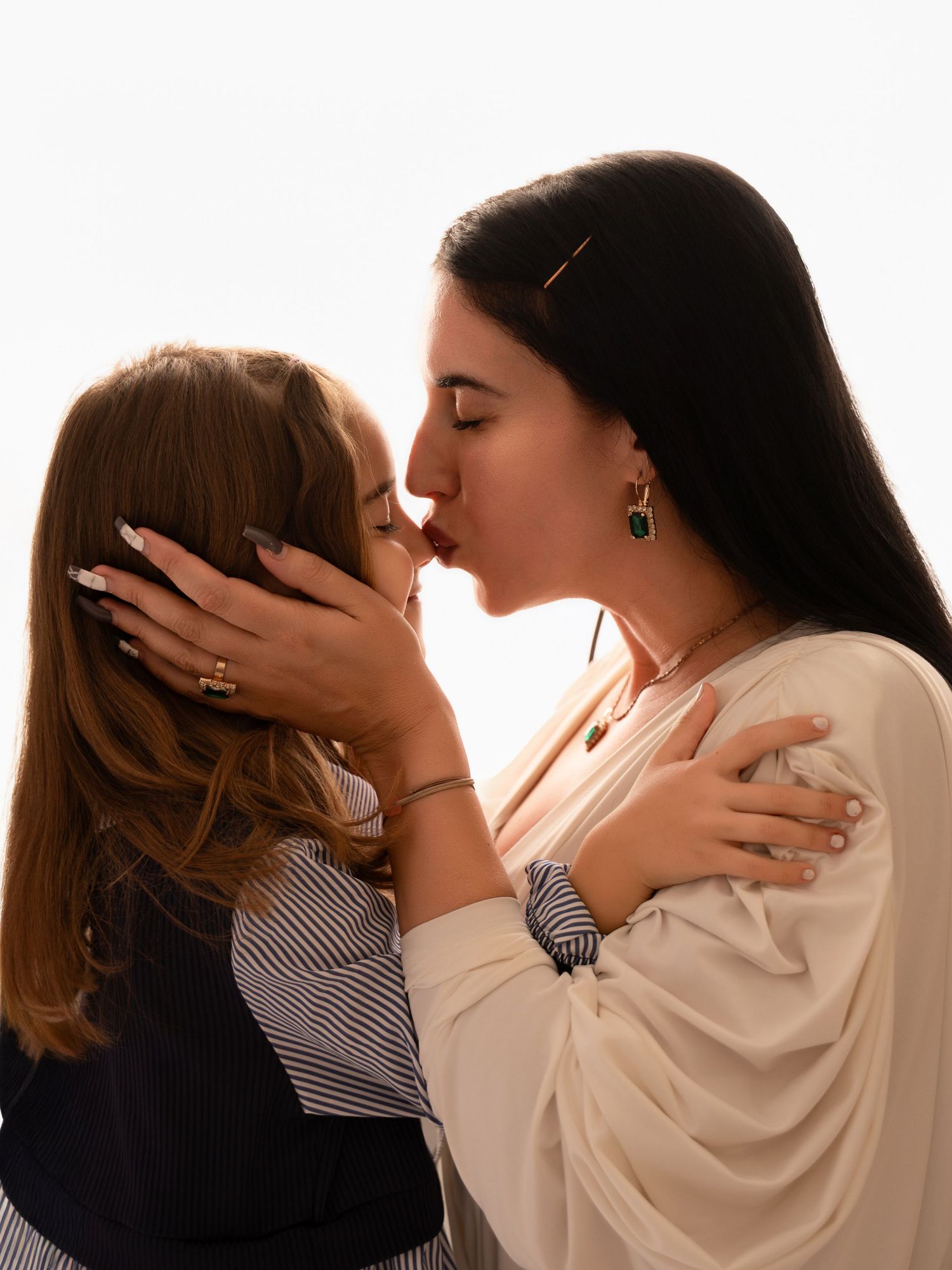 Two women sharing a tender kiss, one with dark hair wearing green earrings, embracing in warm backlit light.