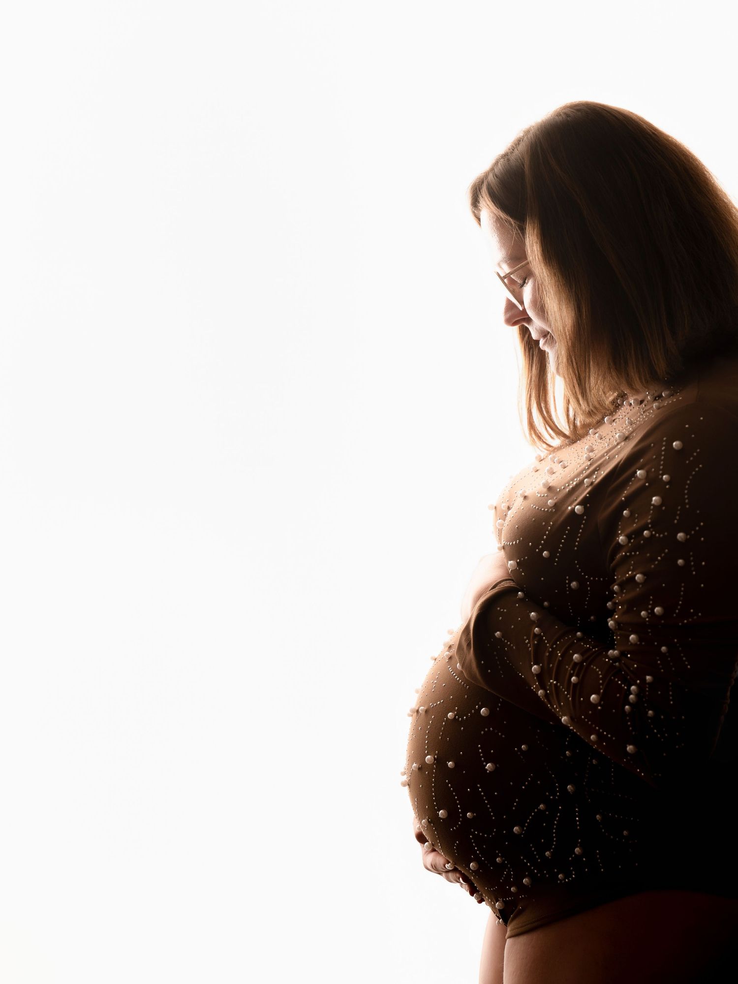 Pregnant woman in beaded gown photographed in profile against bright white background in artistic maternity portrait.