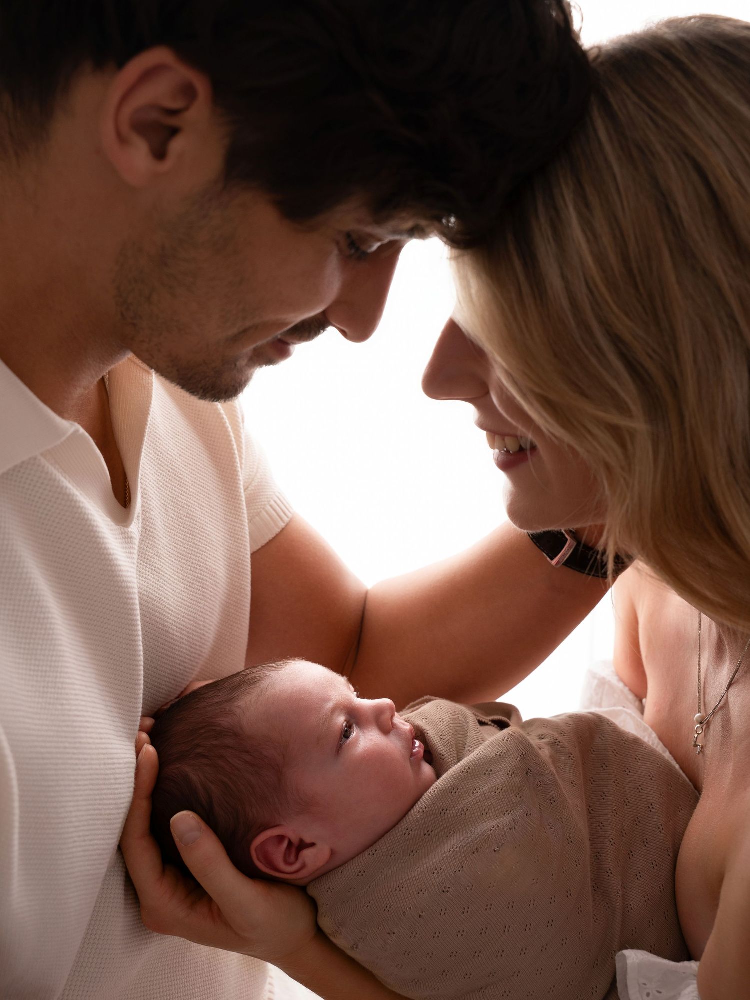 A couple smiles lovingly at their newborn baby wrapped in a blanket, sharing a tender family moment.