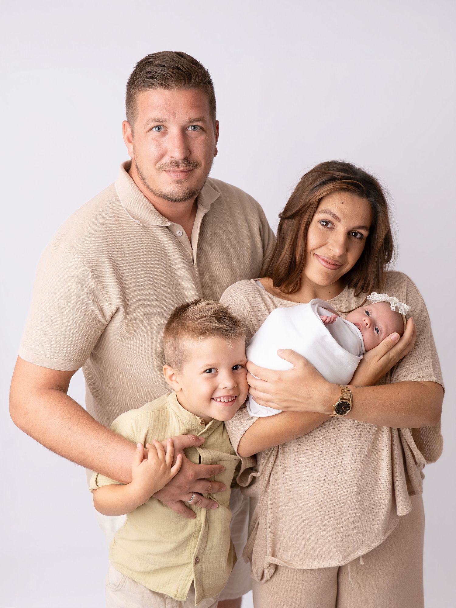 Family portrait of parents with toddler son and newborn baby, wearing coordinated beige outfits on white background.