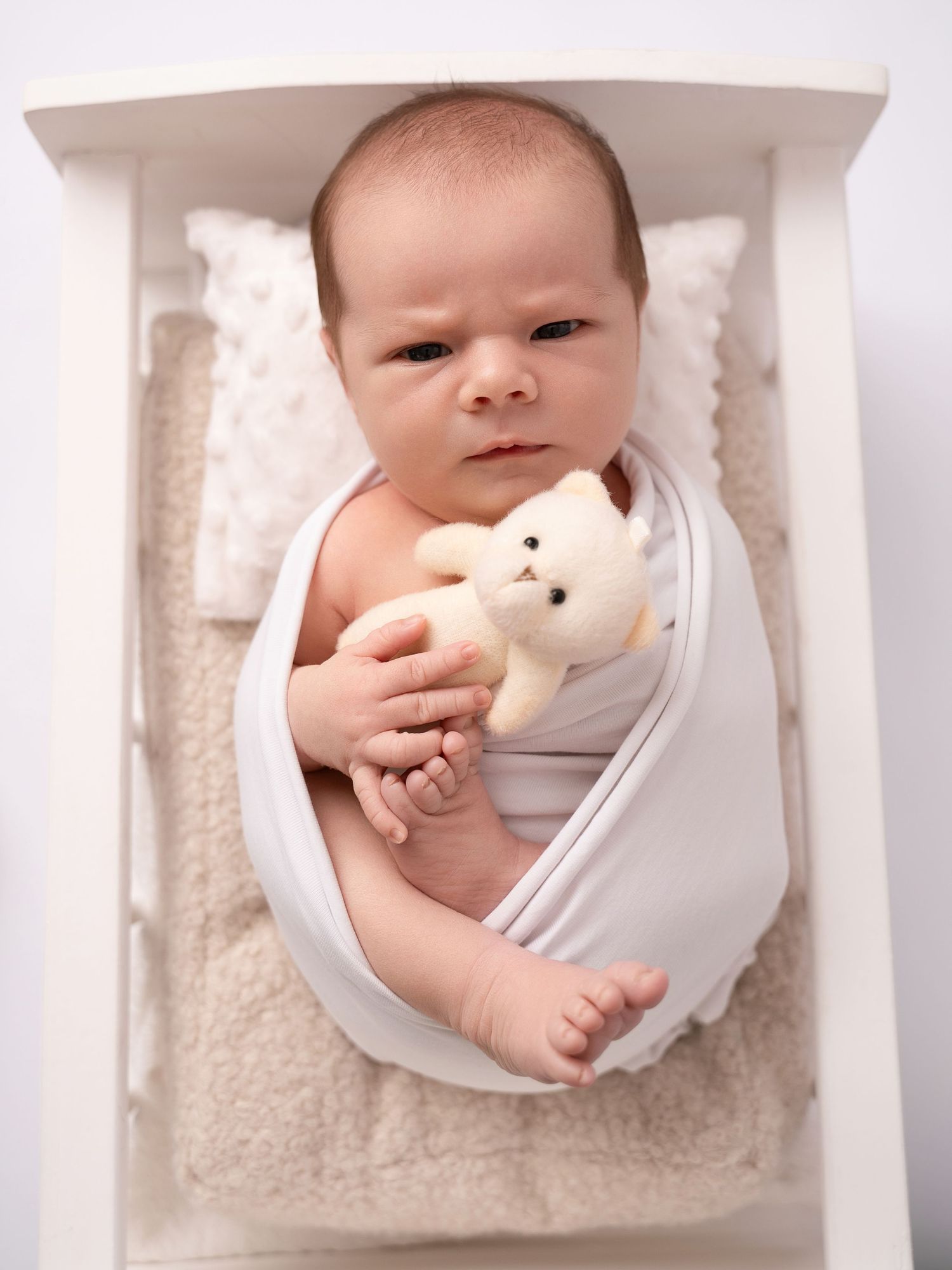 Newborn baby swaddled in white wrap holding a small teddy bear, lying in a white wooden crib on fluffy bedding.
