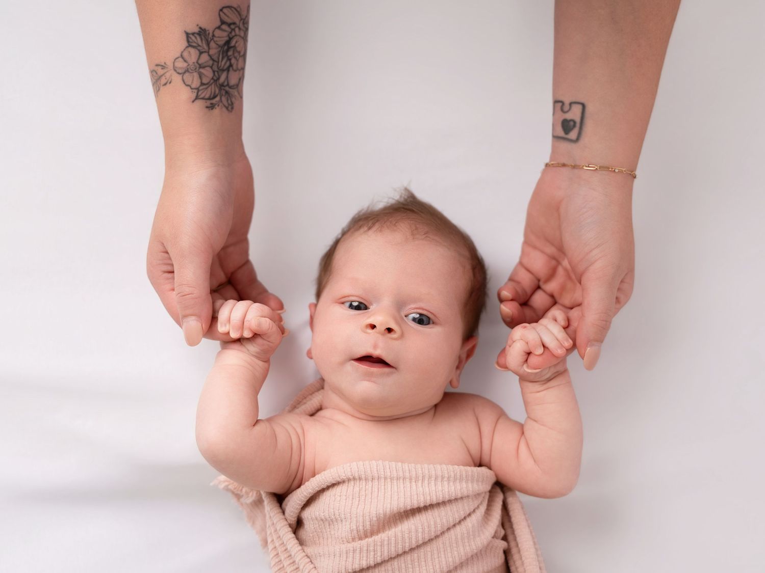 Newborn baby lying on white background held up by tattooed adult hands.
