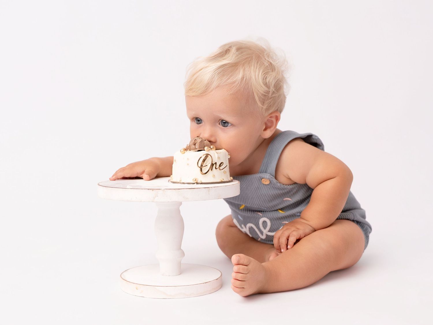 Baby in gray romper eating first birthday cake labeled One during a cake smash photo session.