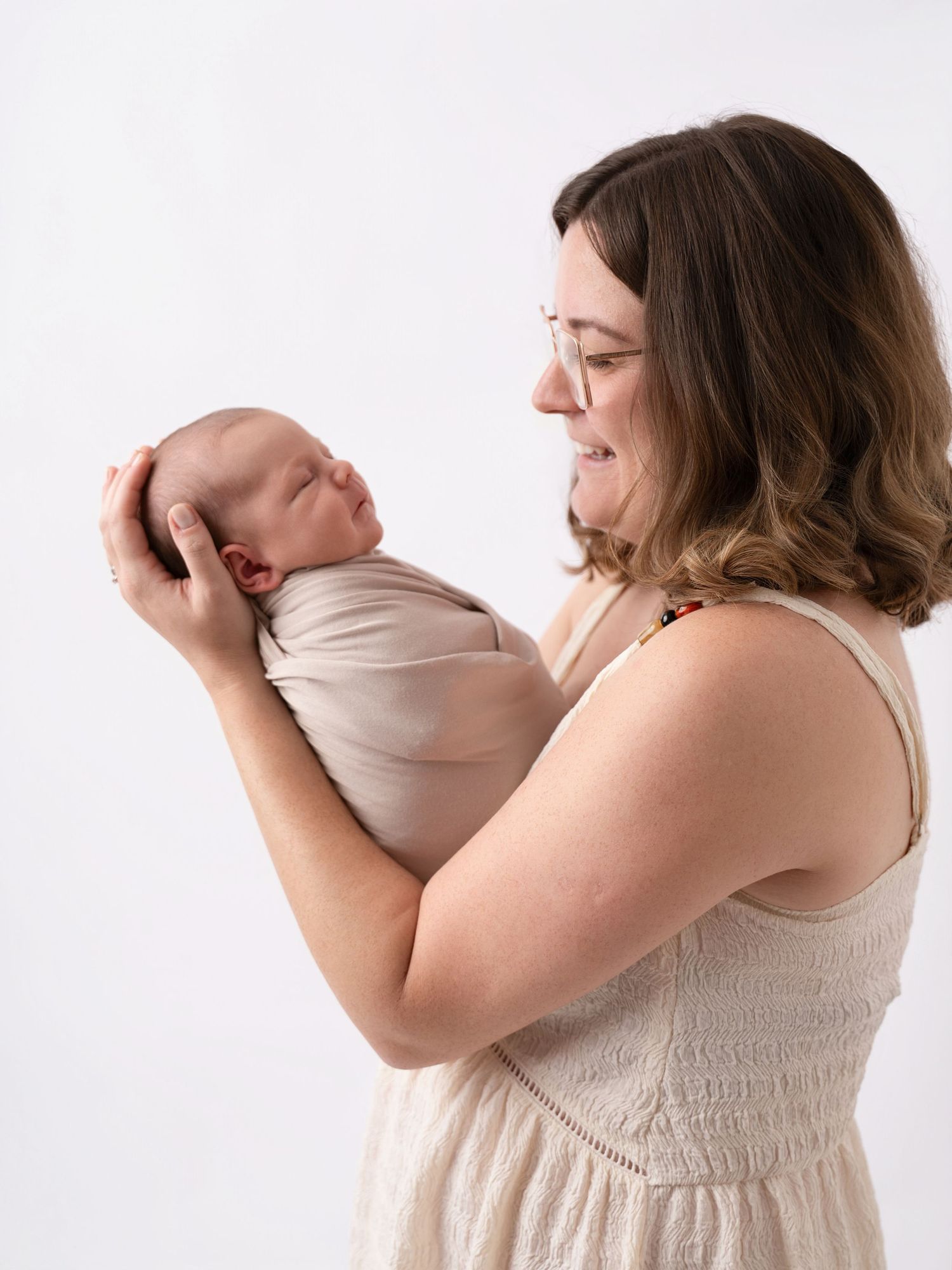 A mother in a cream dress lovingly gazes at her swaddled newborn baby held in her arms.