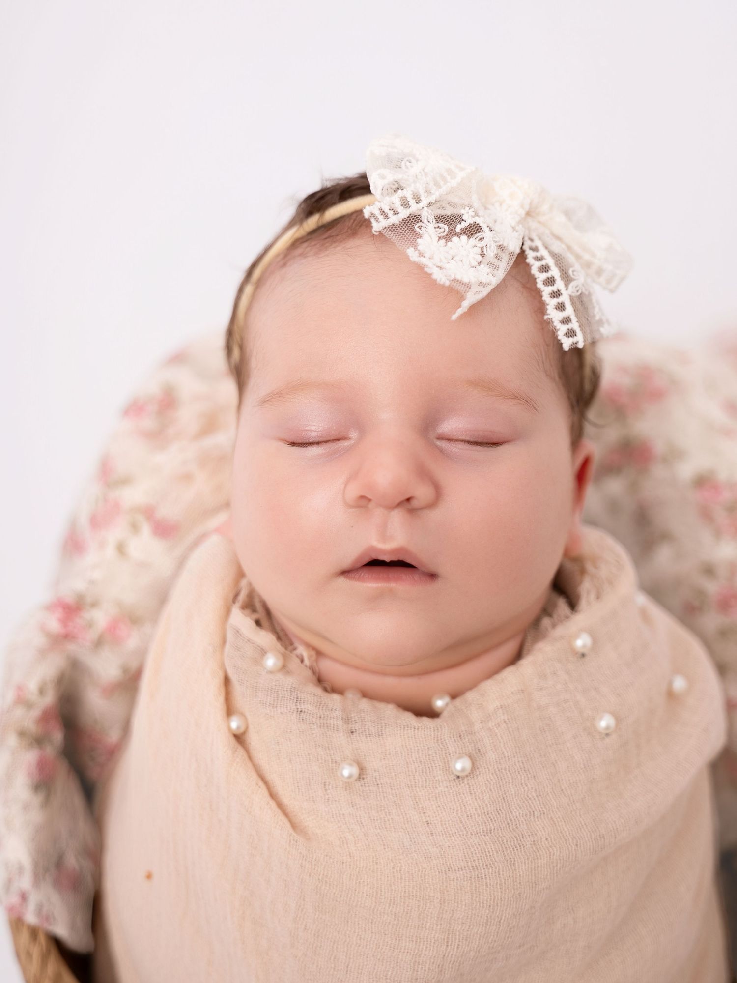 Sleeping newborn baby swaddled in a pearl-adorned wrap, wearing a lace bow headband, photographed on a white background.