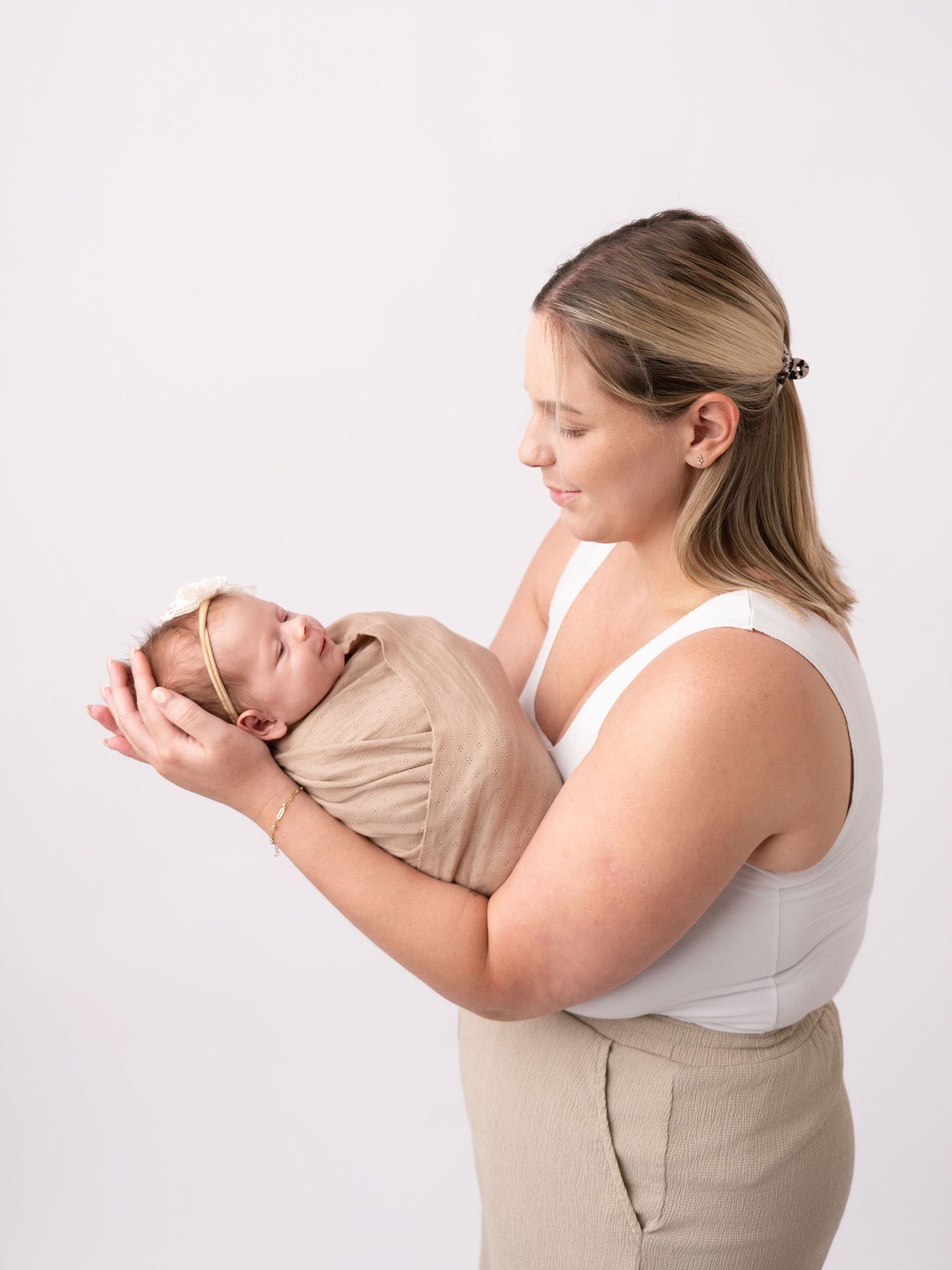 A mother tenderly holds her swaddled newborn baby in a tan wrap with a bow headband against a white background.