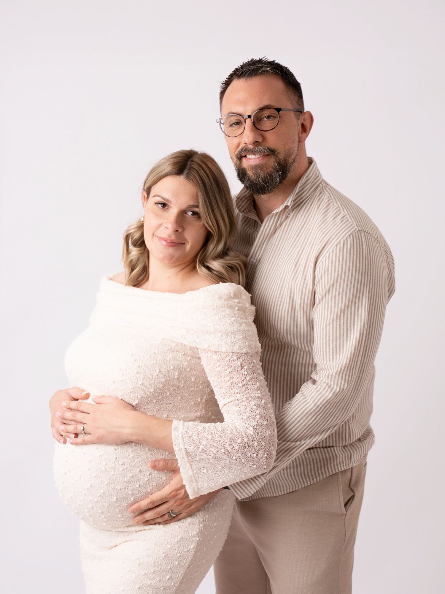 Expecting couple in neutral tones poses tenderly, woman in white lace maternity gown, man in beige shirt, studio photo.