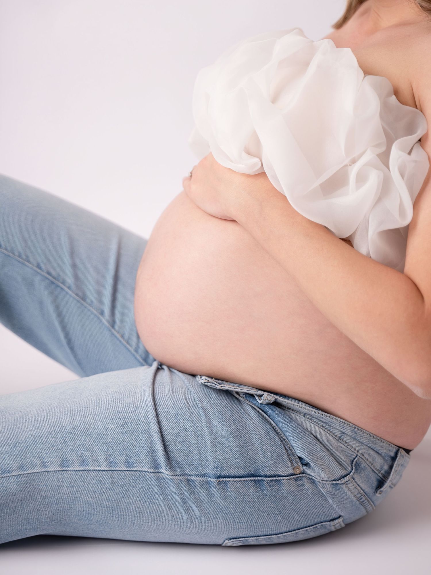 Pregnant woman sitting on white background, wearing jeans and white ruffled top, cradling her bare baby bump.