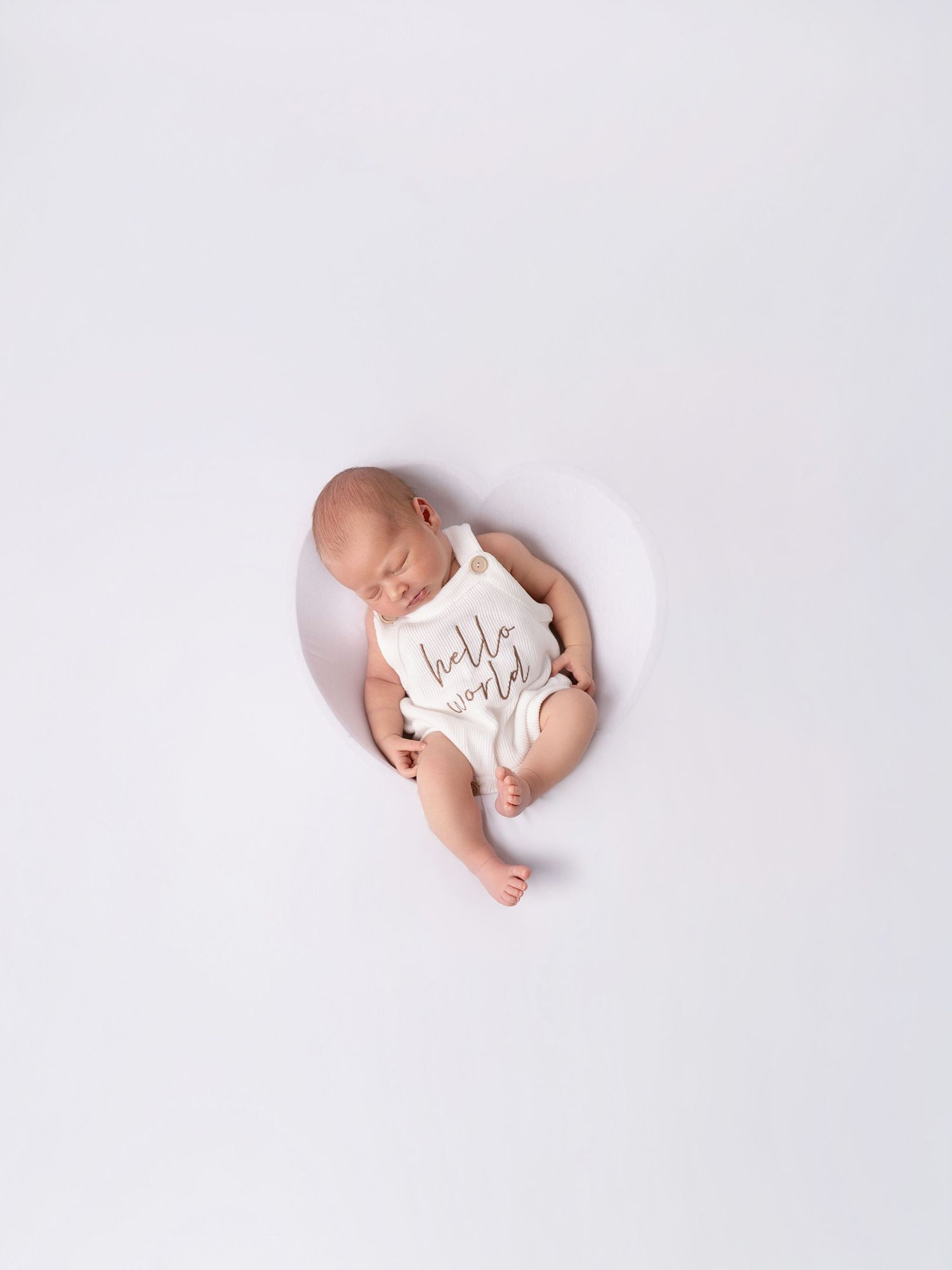 Newborn baby sleeping peacefully in a white onesie that reads hello world, photographed from above on white background.