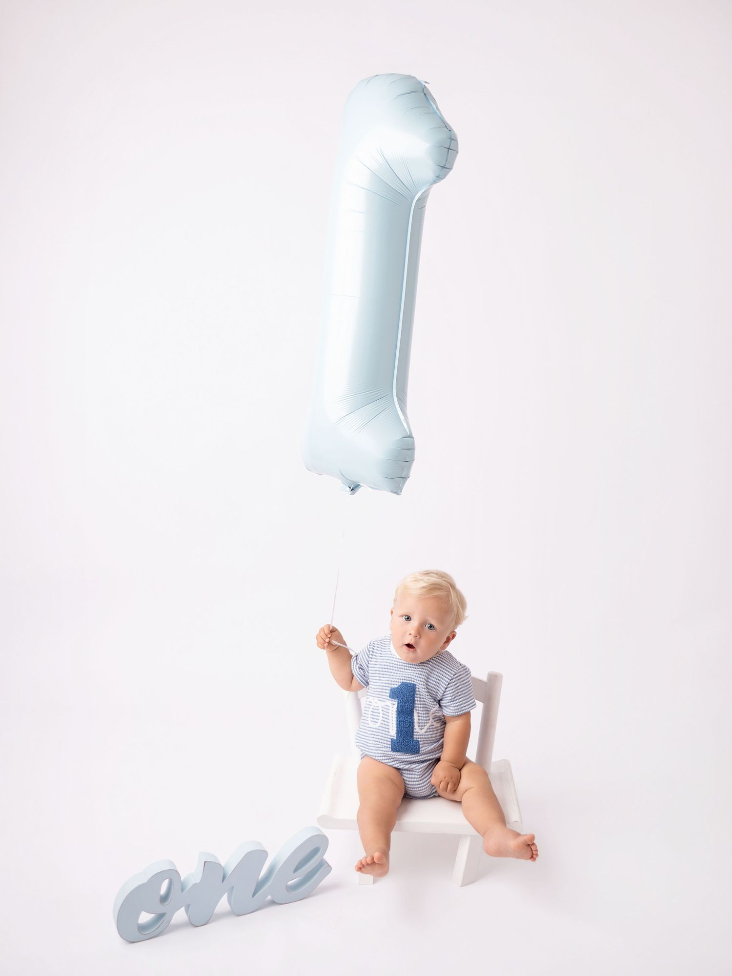 Baby boy in a '1' shirt sits on white chair with blue balloon and 'one' sign for first birthday photo.