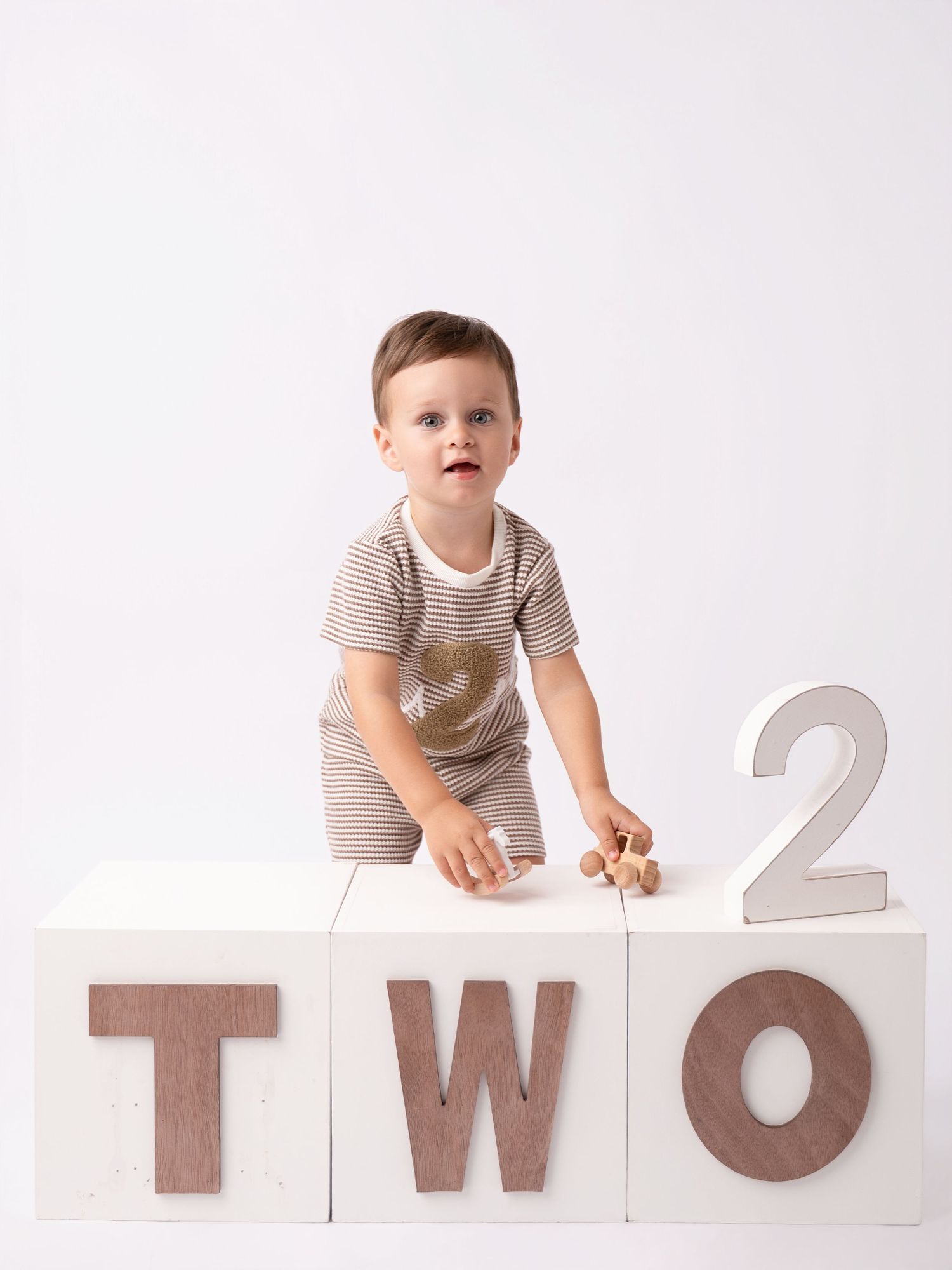 Toddler boy in striped outfit standing behind white blocks spelling TWO for a second birthday photo.