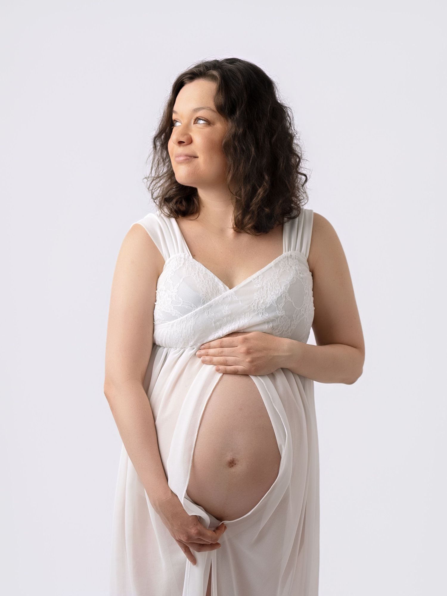 Pregnant woman in white dress cradling her bare baby bump during a maternity photo session.