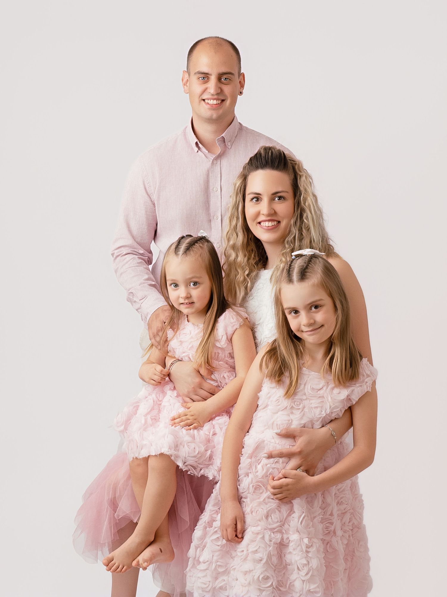 A family of four in coordinating pink outfits poses together for a studio portrait against a light background.