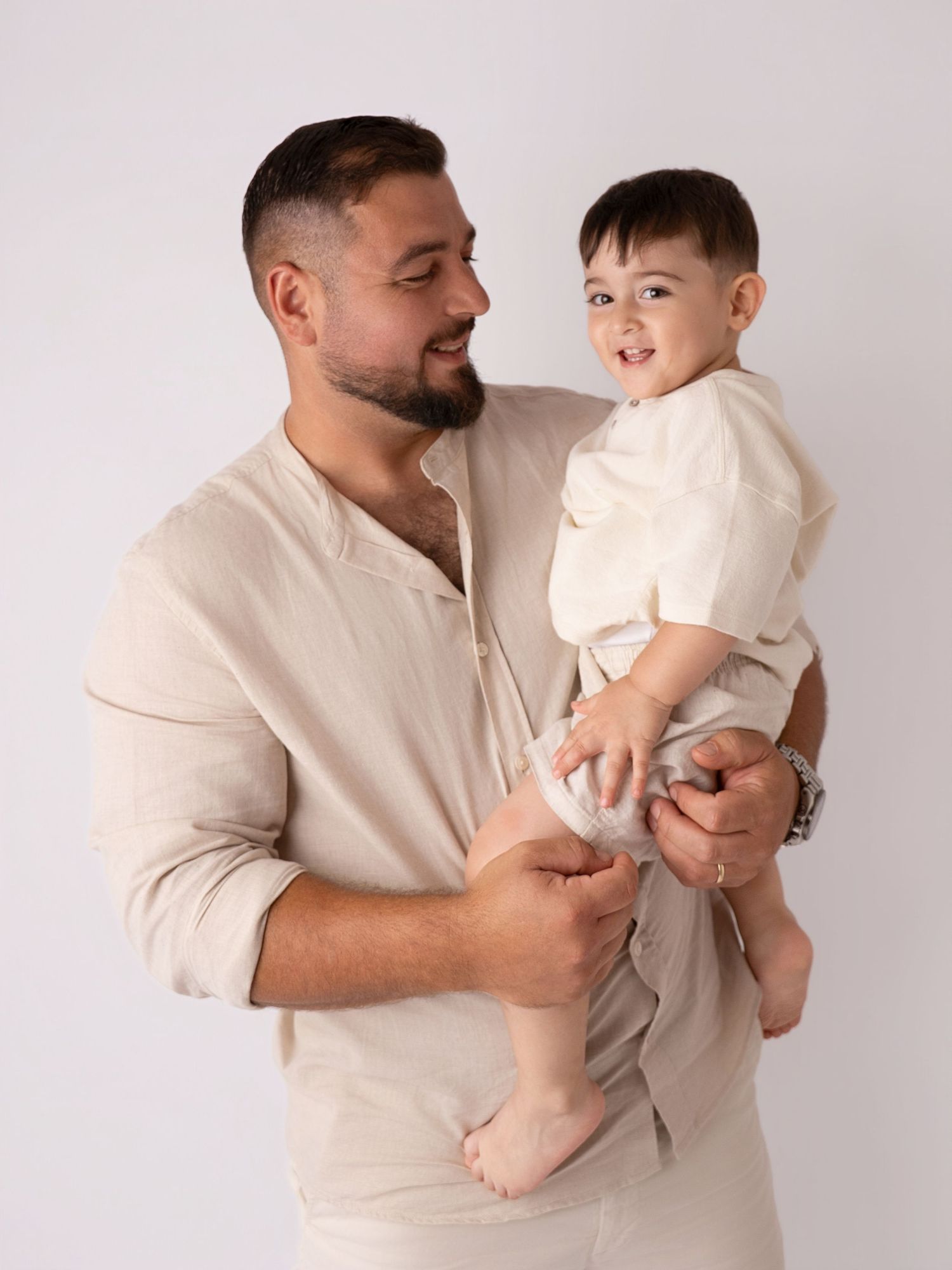 Bearded father in beige linen shirt holds smiling baby boy in a bright studio portrait.