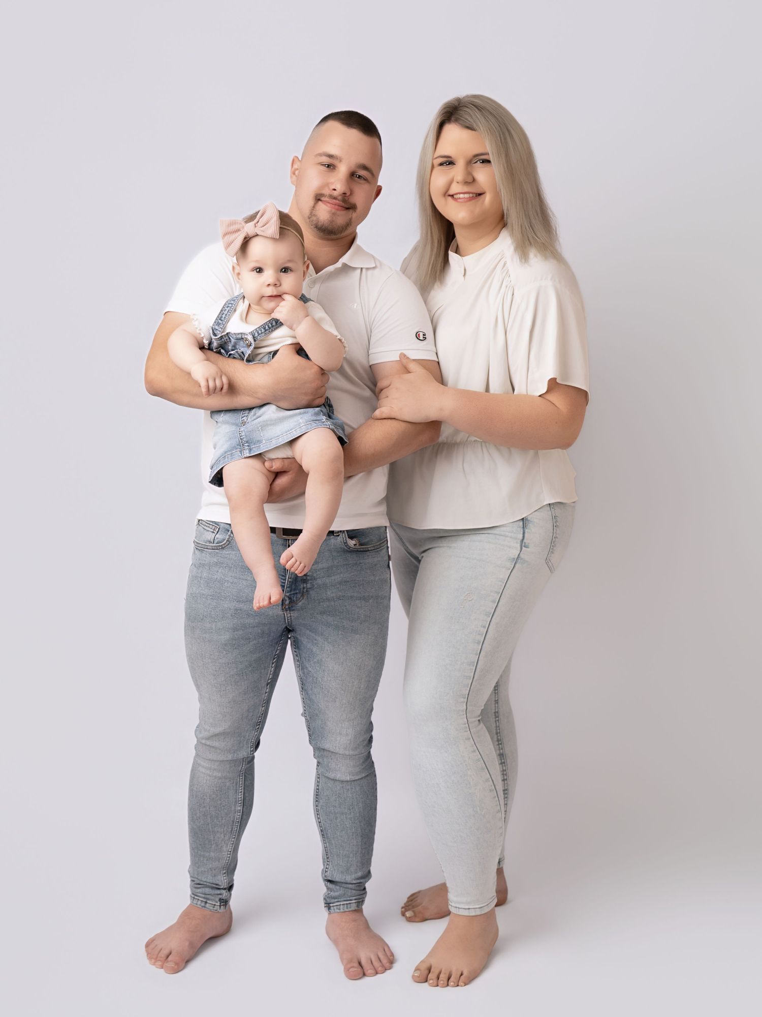 A young family poses together, parents in white tops and gray jeans holding their smiling baby girl in a studio.
