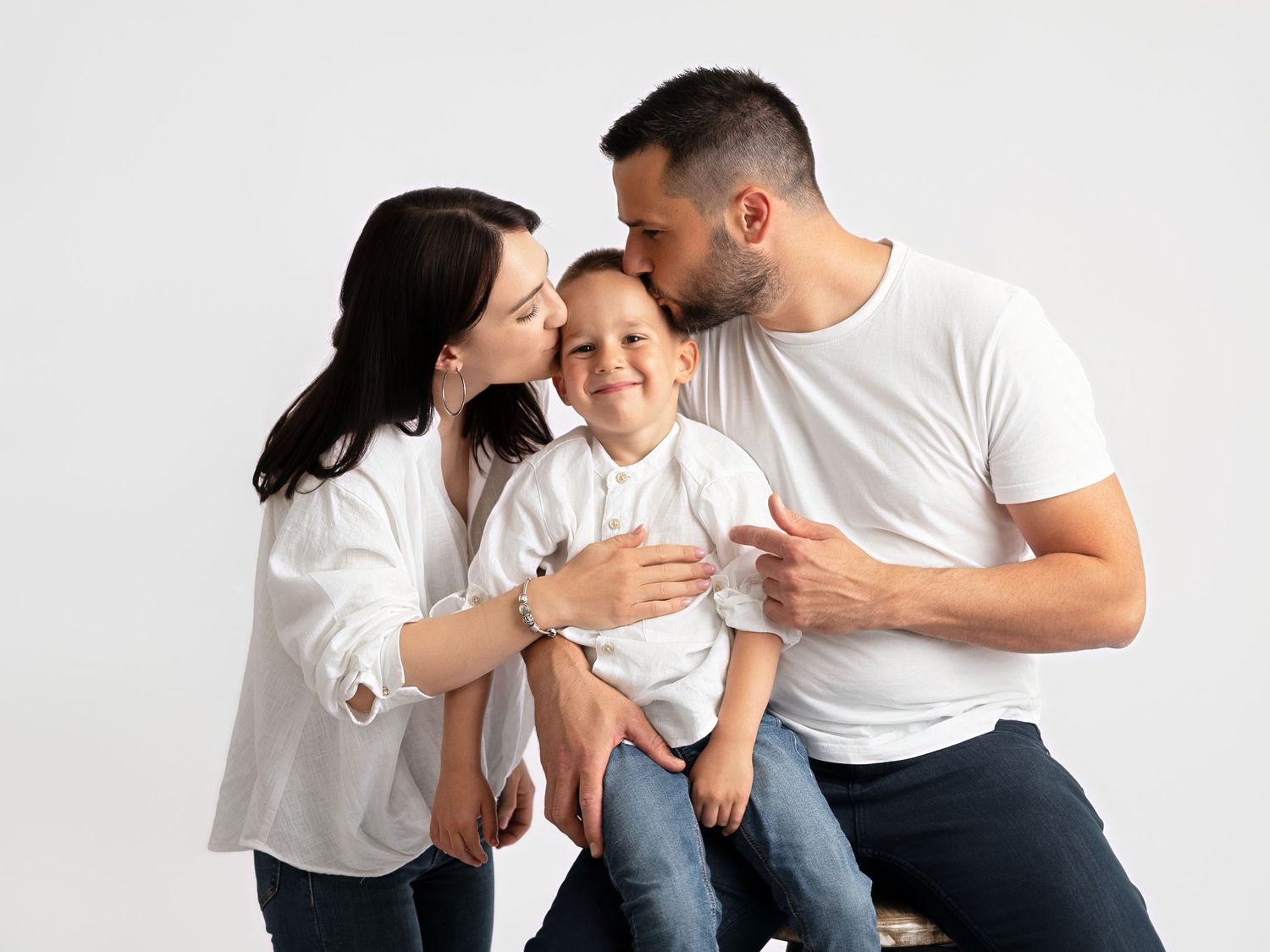 A happy family portrait with parents kissing their young son on the cheeks against a white background.