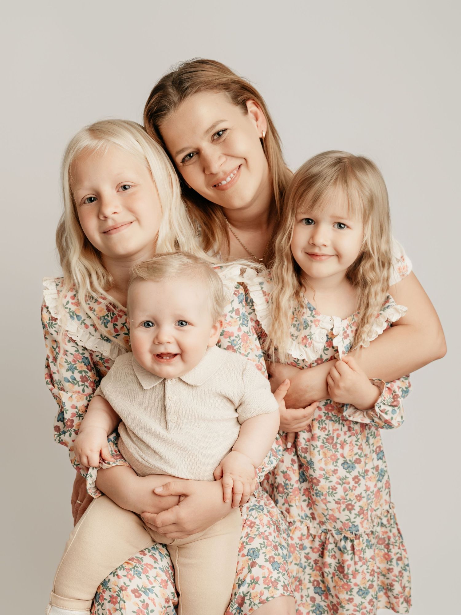 Smiling mother with three young blonde children in floral outfits posing for family studio portrait.