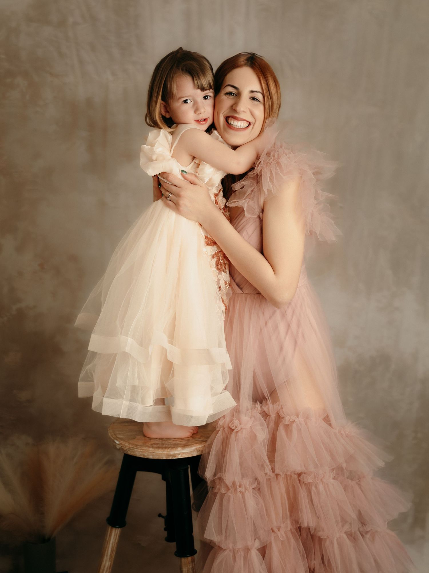 A smiling mother in a pink ruffled tulle gown hugs her young daughter in a white tulle dress during a studio portrait session.