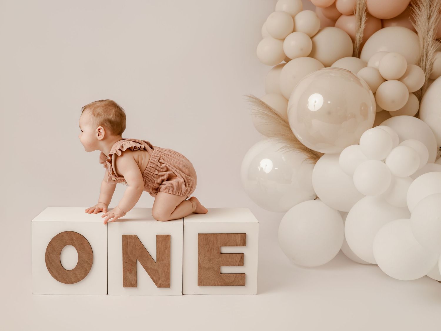 Baby in brown romper crawls on wooden ONE blocks surrounded by neutral balloons for a first birthday cake smash photo.