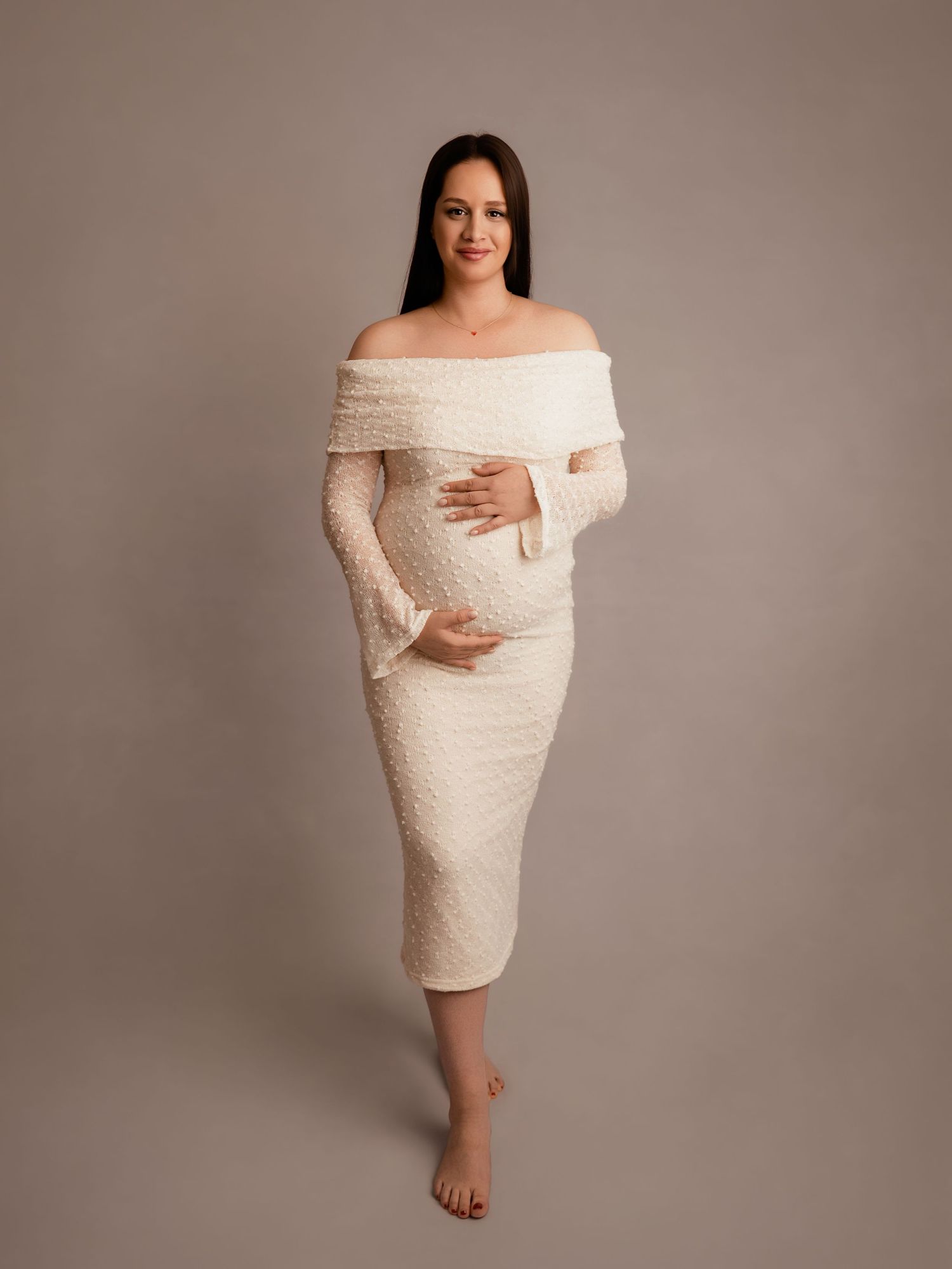 Pregnant woman in elegant off-shoulder cream knit dress posing gracefully in a professional maternity photo studio.
