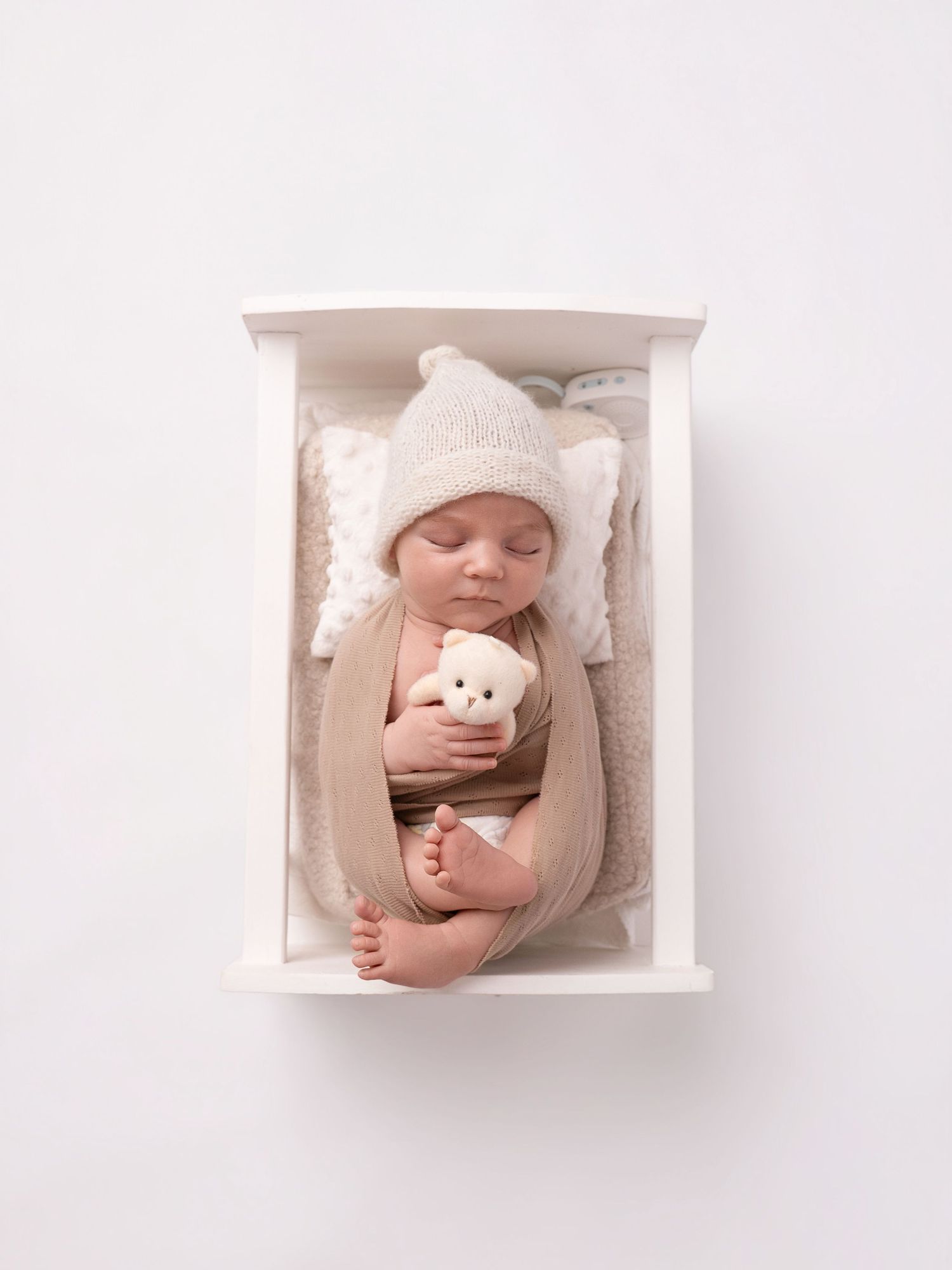 Newborn baby swaddled in beige wrap, wearing knit hat, holding white bear toy, sleeping peacefully in white wooden box.