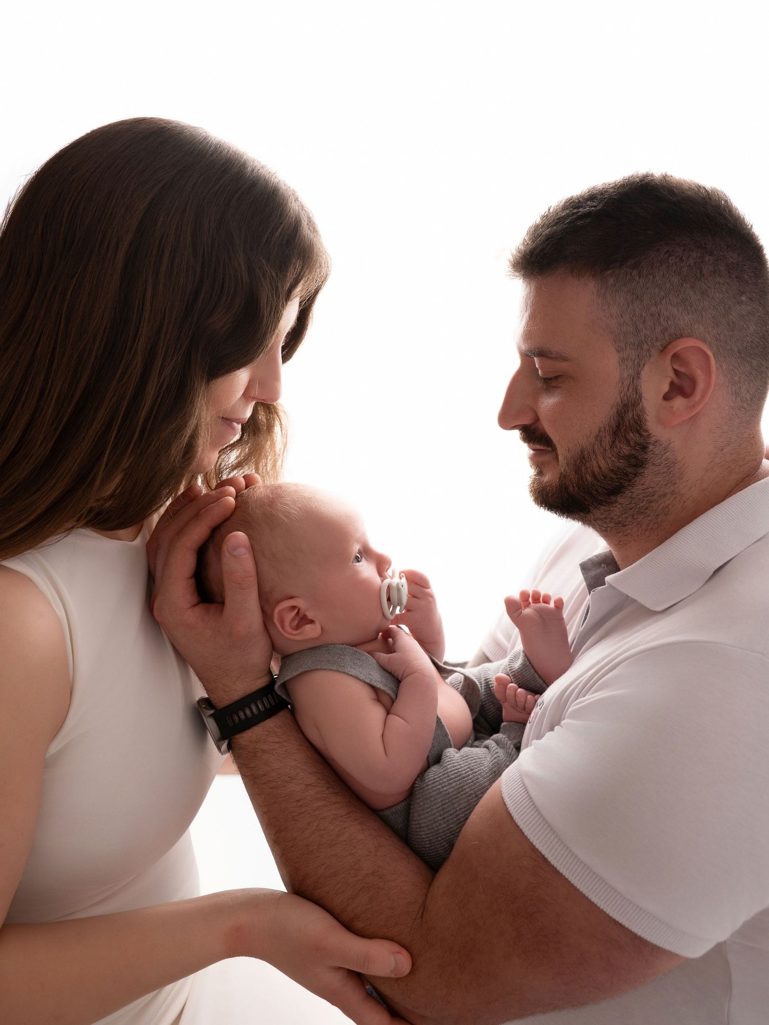 A couple lovingly gazes at their newborn baby, cradled in their arms against a bright white background.
