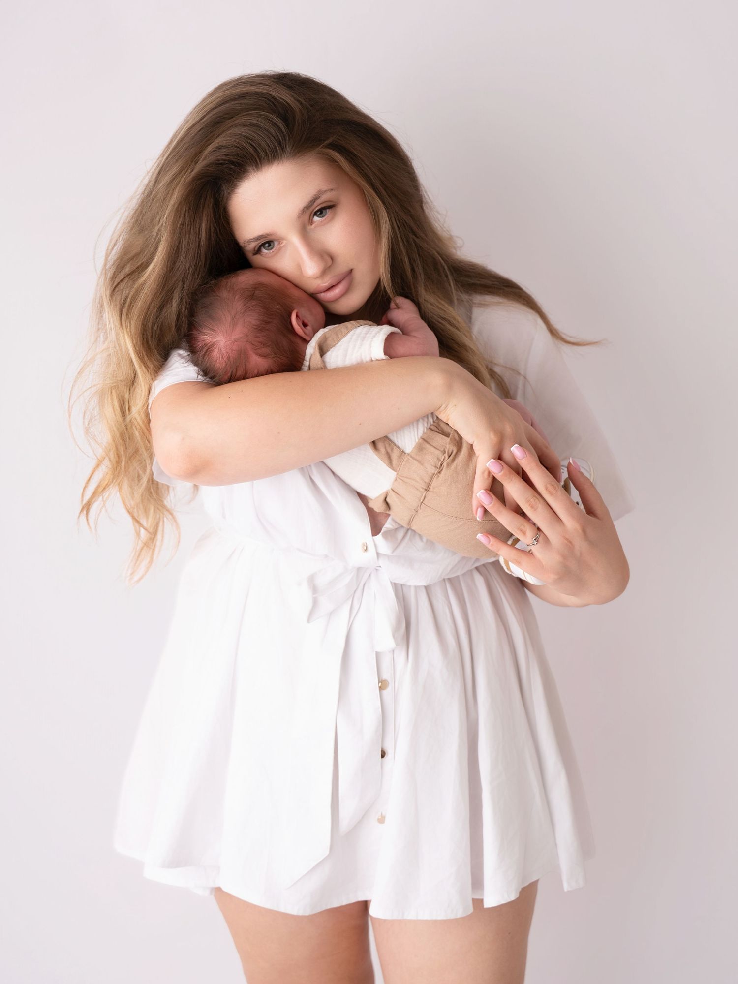 Young mother in white dress tenderly holding sleeping newborn baby against light background.