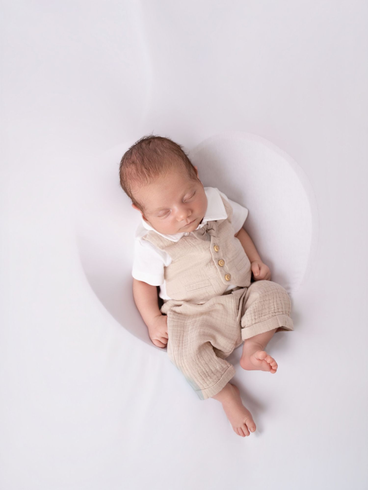 Sleeping newborn baby boy wearing a beige linen vest and pants outfit on a white heart-shaped backdrop.
